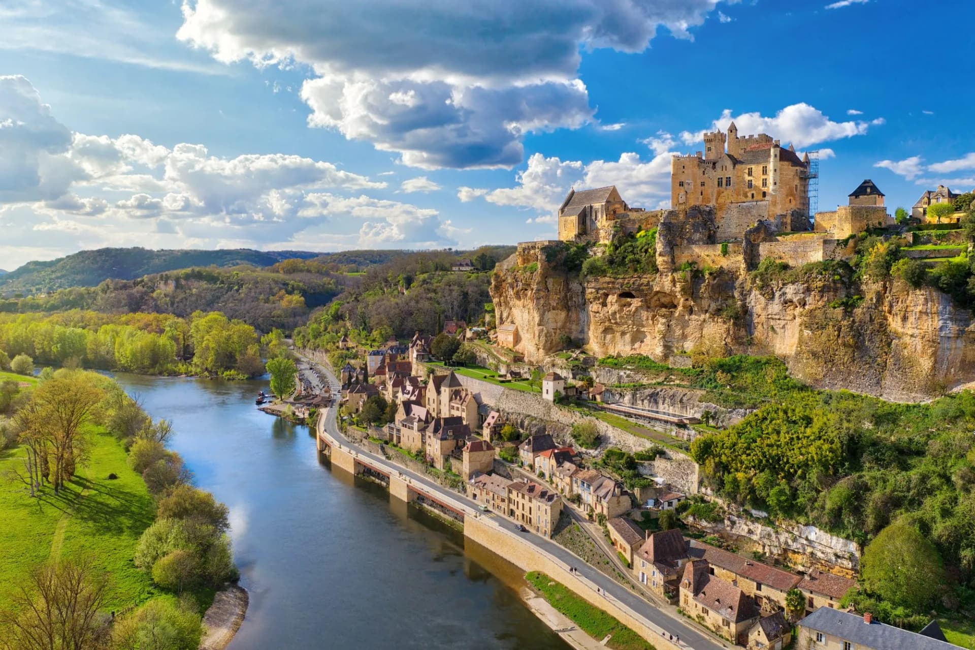 Sarlat castle perched on a cliff above a river and historic village under a blue sky.
