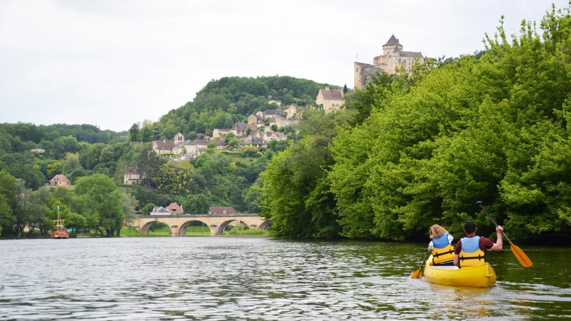 Canoeing on river past hillside village with stone bridge and castle ruins