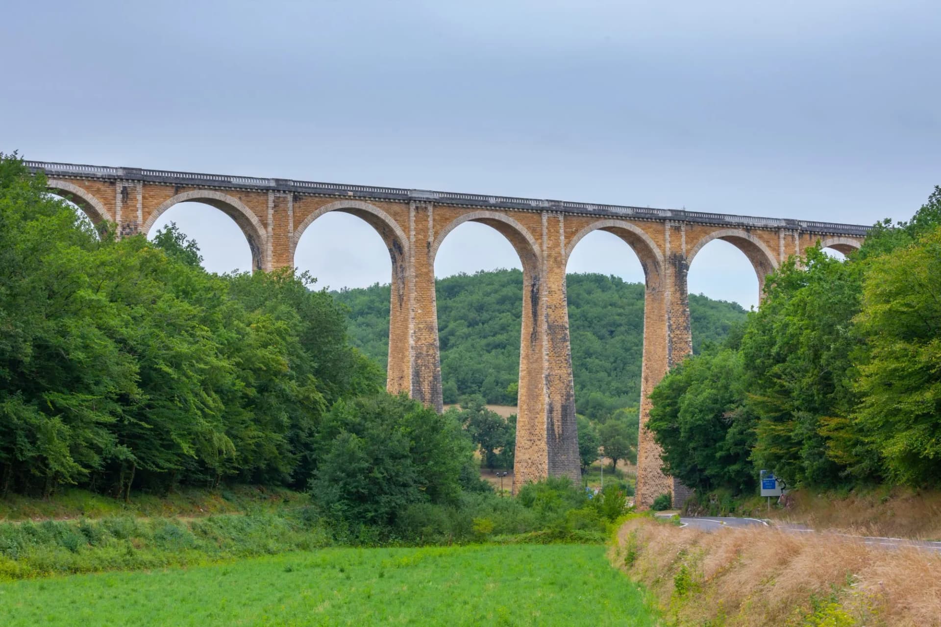 Stone arch viaduct rising above lush green forest and a roadside field under a gray sky.