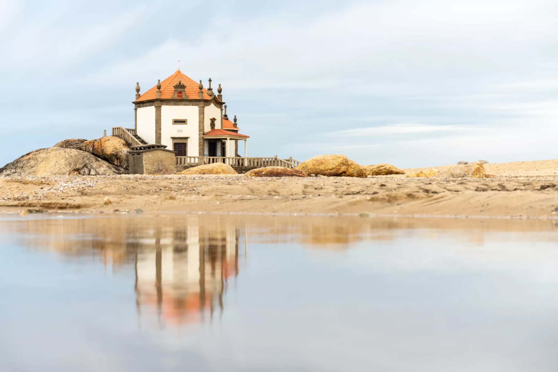 Senhor da Pedra Chapel reflected in tidal pool on sandy beach with large rocks.
