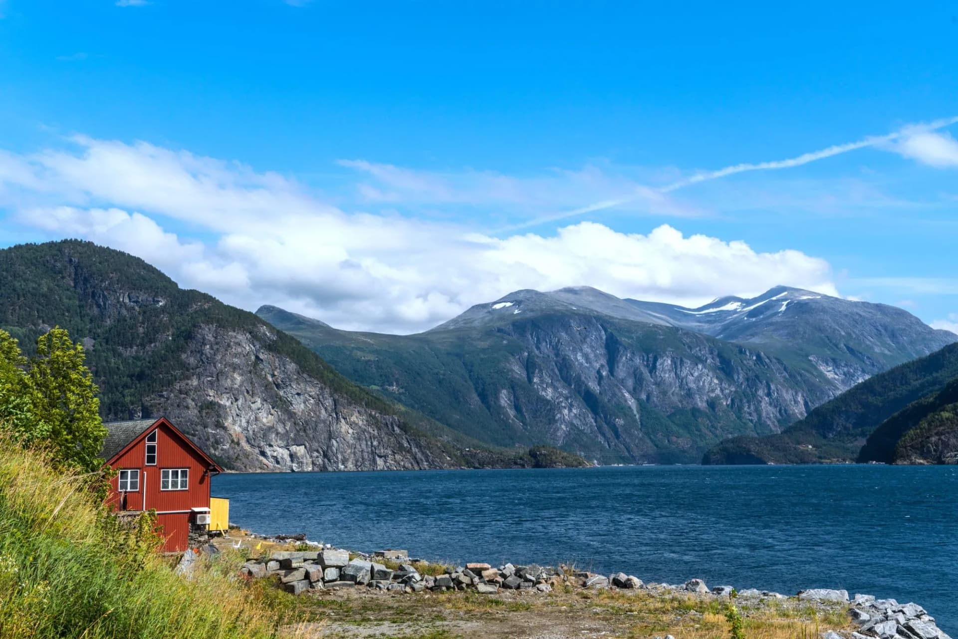 Red house by Storfjorden with steep, forested mountains under a blue sky.
