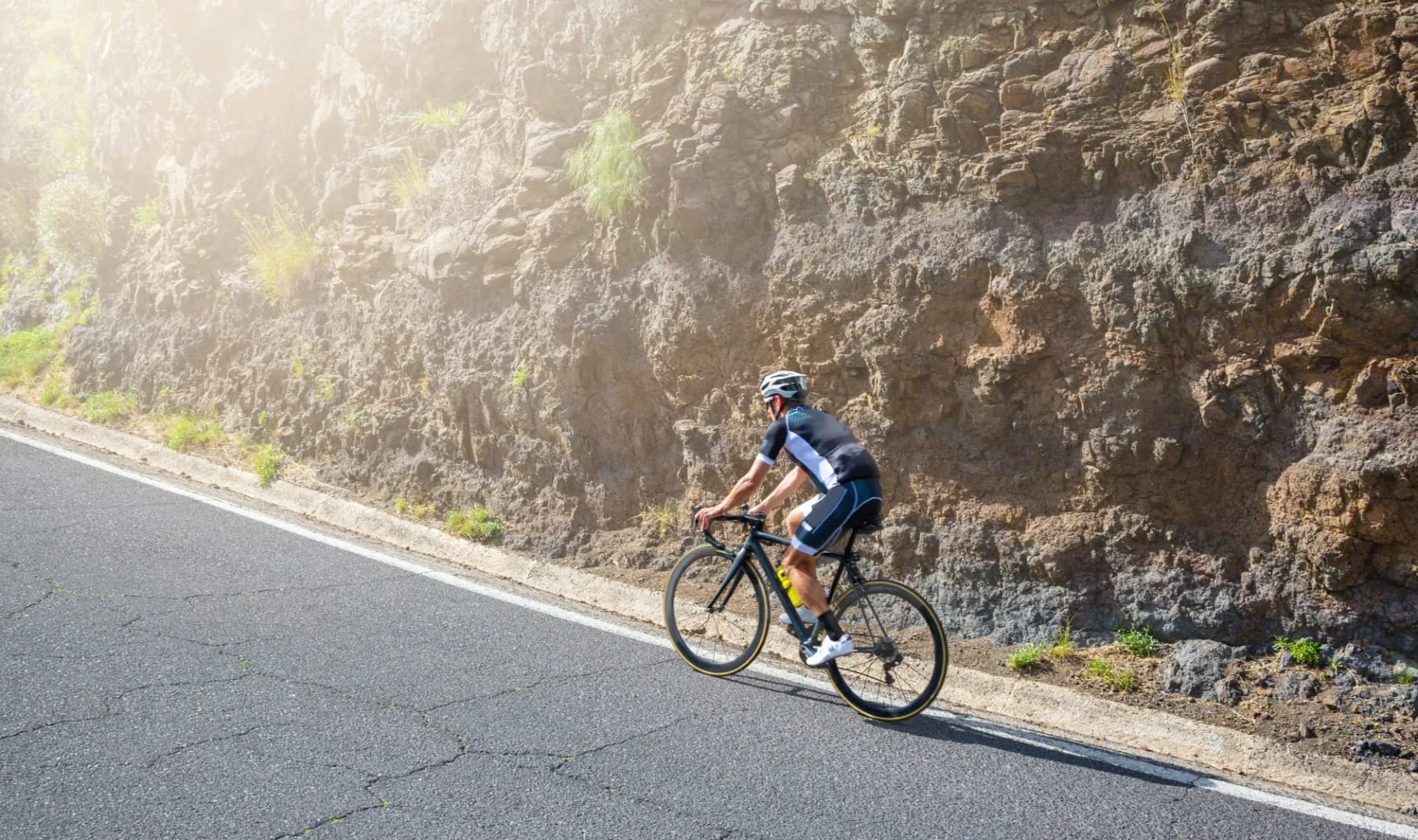Road cyclist riding uphill next to a steep, sunlit rocky embankment.