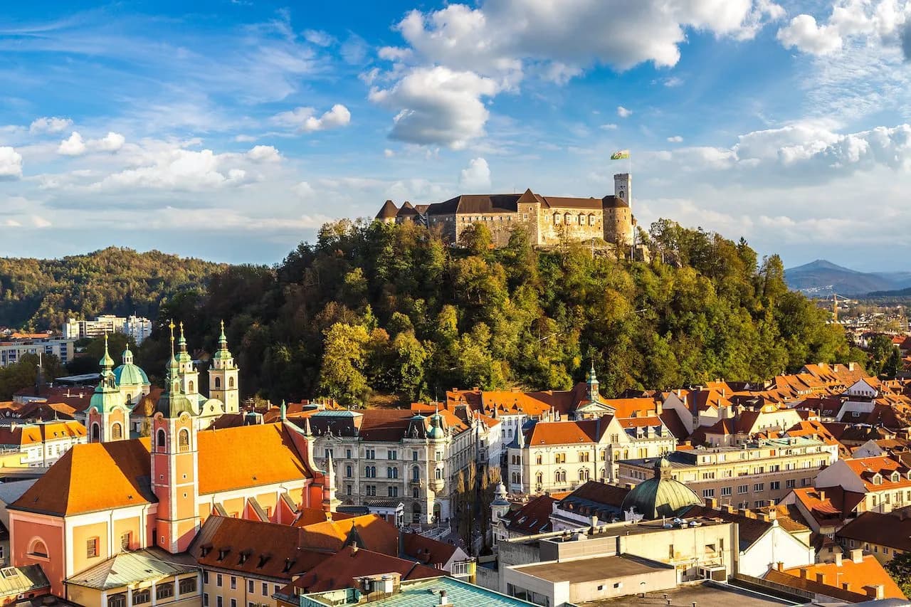 Ljubljana Castle above old town with orange roofs and green church spires under blue sky.