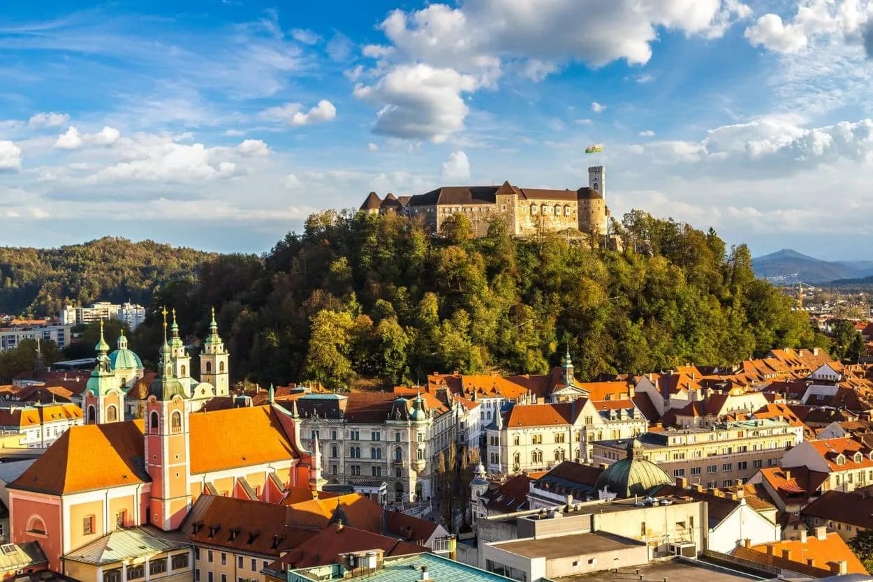 Ljubljana Castle atop a wooded hill overlooking city rooftops and a pink church.