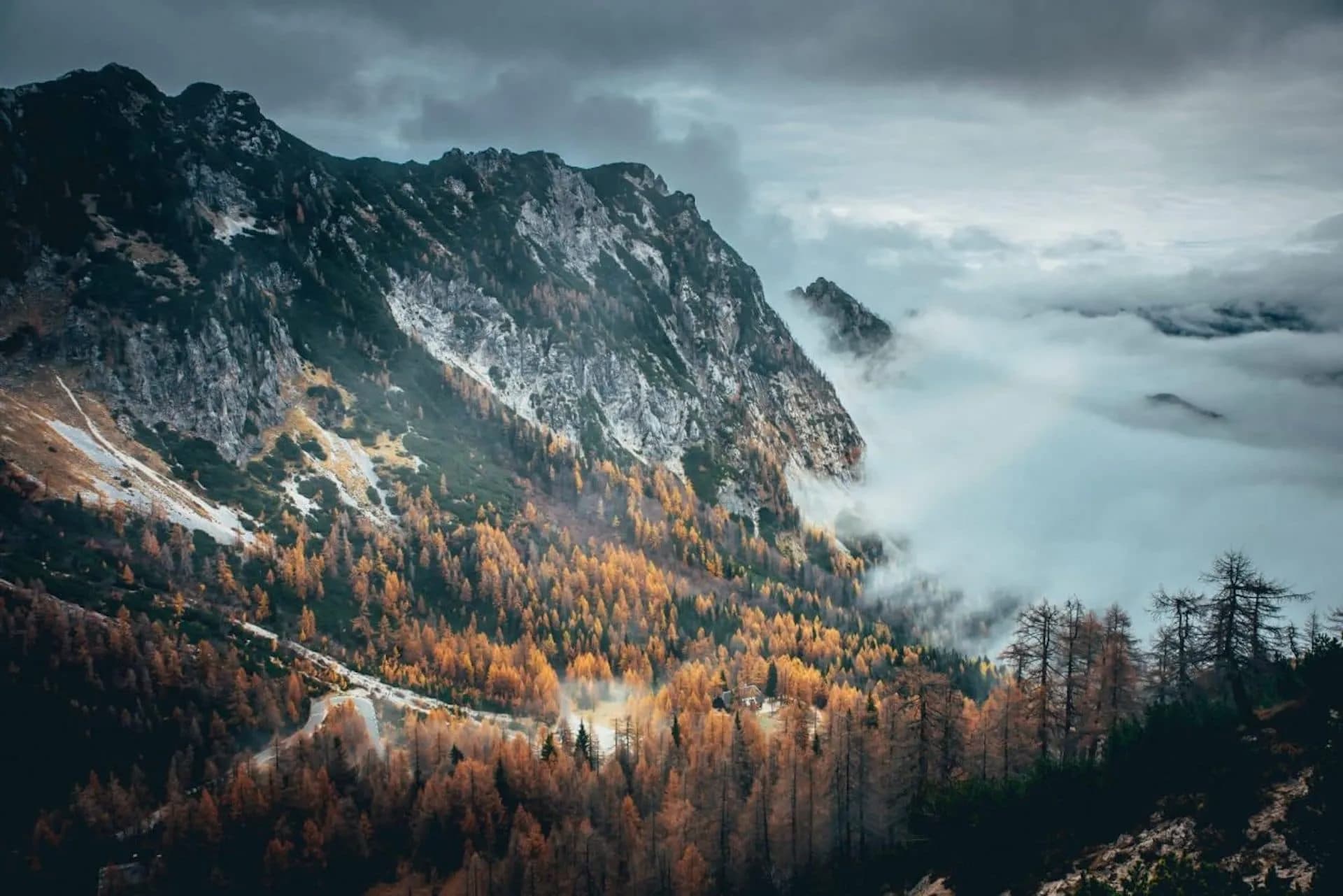 Winding mountain road through autumn forest below rocky peaks shrouded in fog at Vršič Pass, Slovenia.