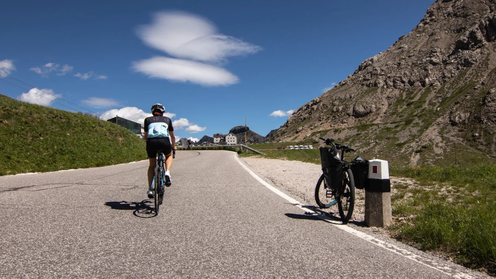 Cyclist riding up a paved mountain pass road toward buildings, Pordoi Pass, Sella Ronda.