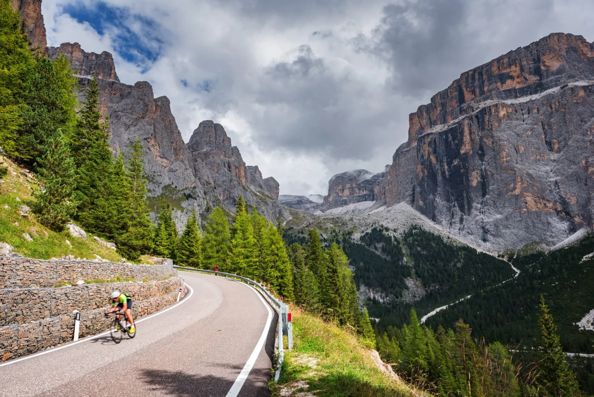 Road cyclist climbing mountain pass with steep rock faces and pine trees in Dolomites