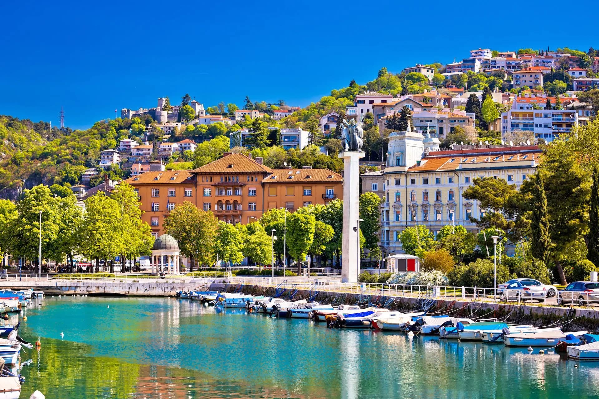Boats moored in turquoise harbor water with historic buildings and a hillside town in Rijeka.