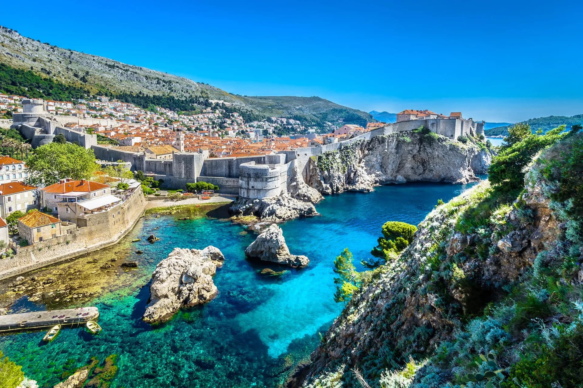 Dubrovnik city walls and terracotta roofs overlooking turquoise Adriatic Sea cove under clear blue sky.
