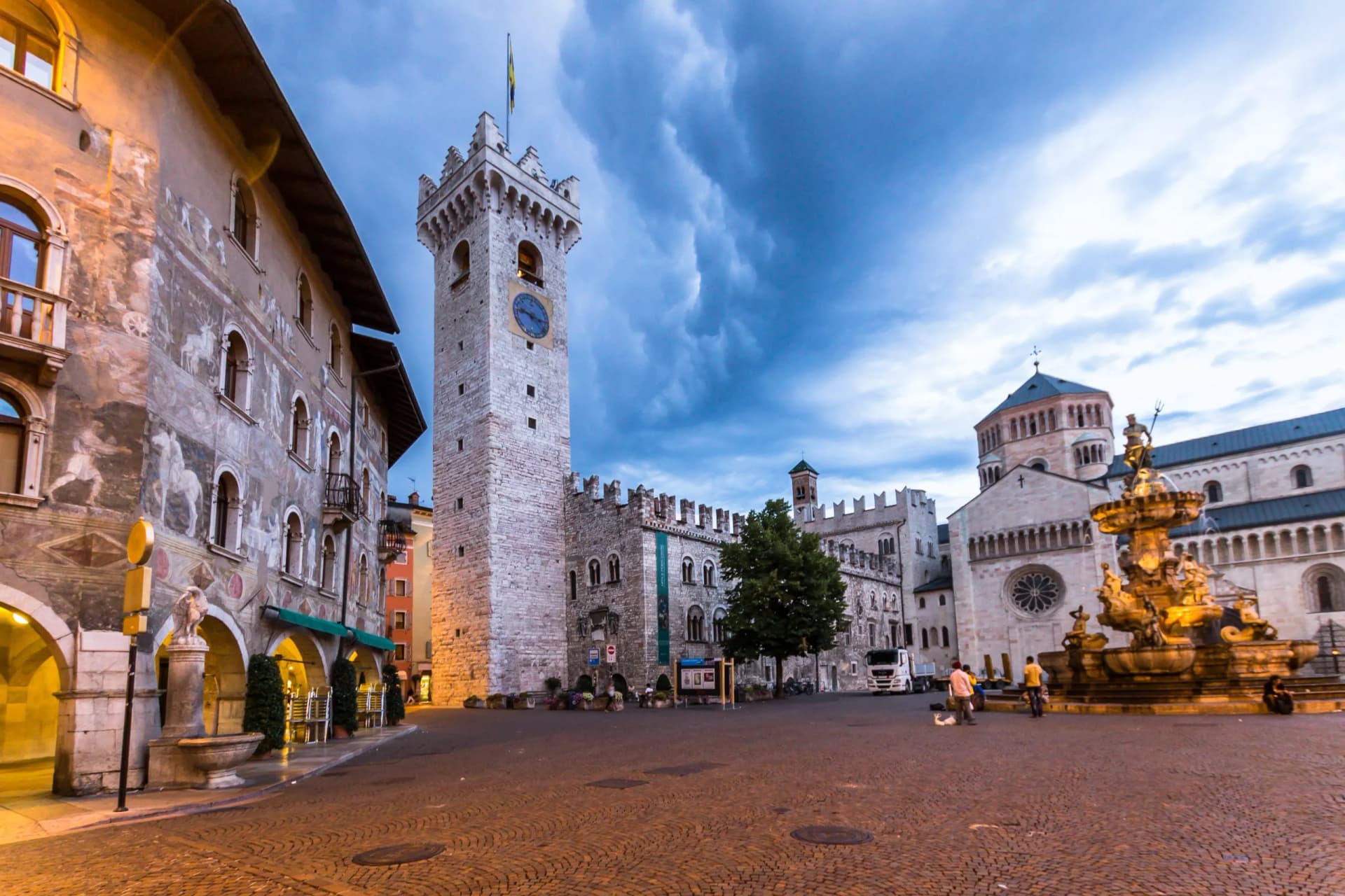 Piazza Duomo in Trento with clock tower, cathedral, and Neptune Fountain under dark clouds.