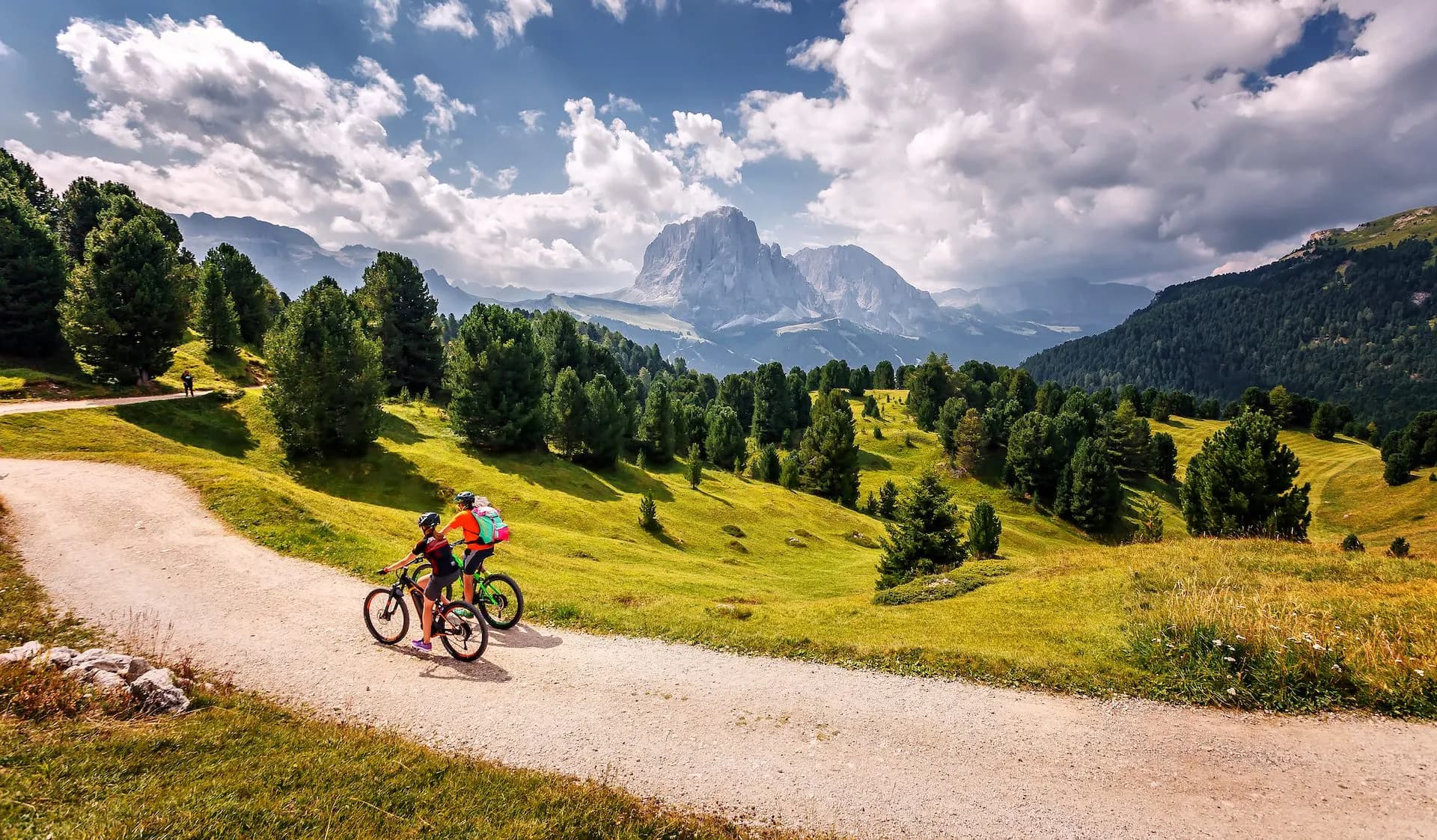 Mountain biking on dirt road with Dolomites Alps peaks and green meadows in background