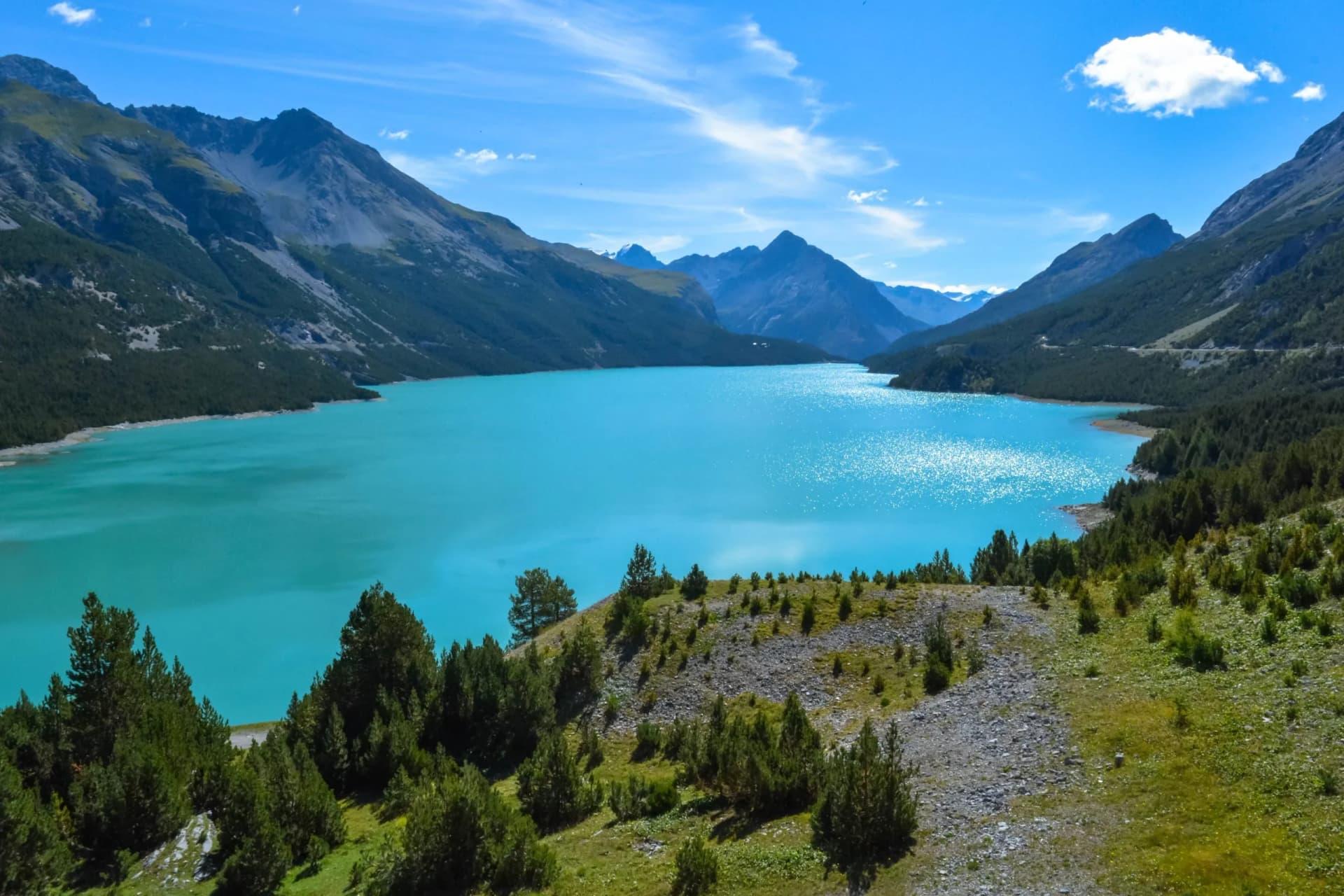 Alpine lake with bright turquoise water surrounded by steep, forested mountains under a blue sky.