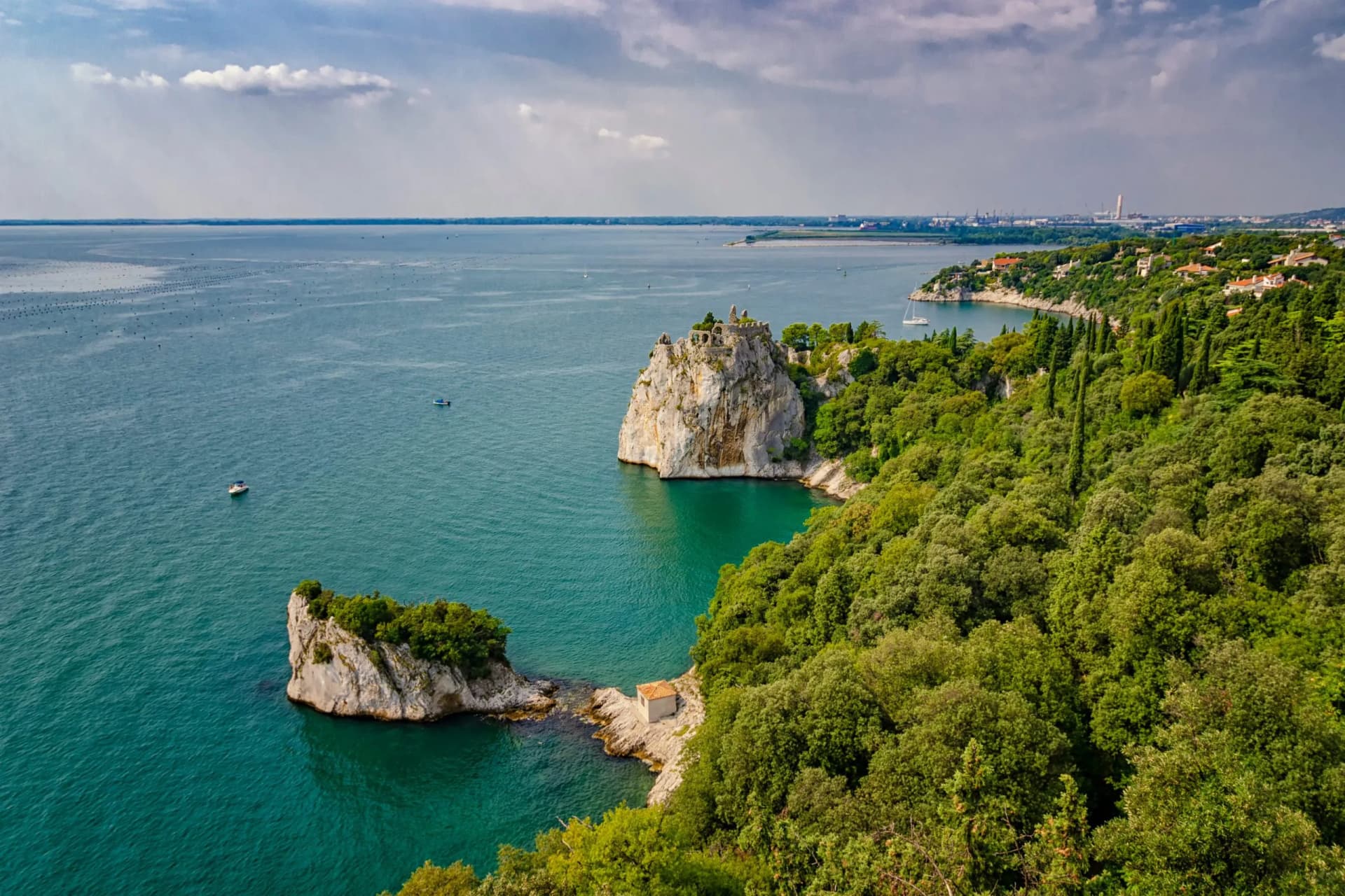 Rocky outcrop with castle ruins overlooking turquoise Gulf of Trieste waters and lush green coastline.