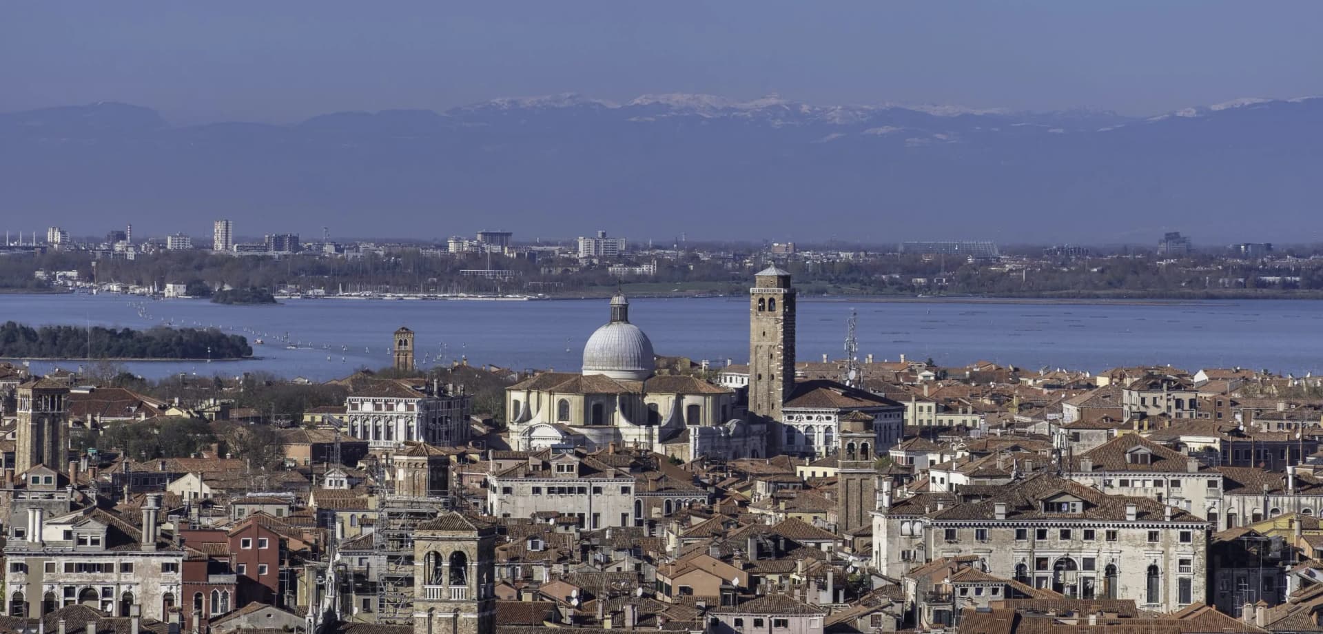 Rooftops of Venice with a large dome church, lagoon, and snow-capped mountains in the distance.