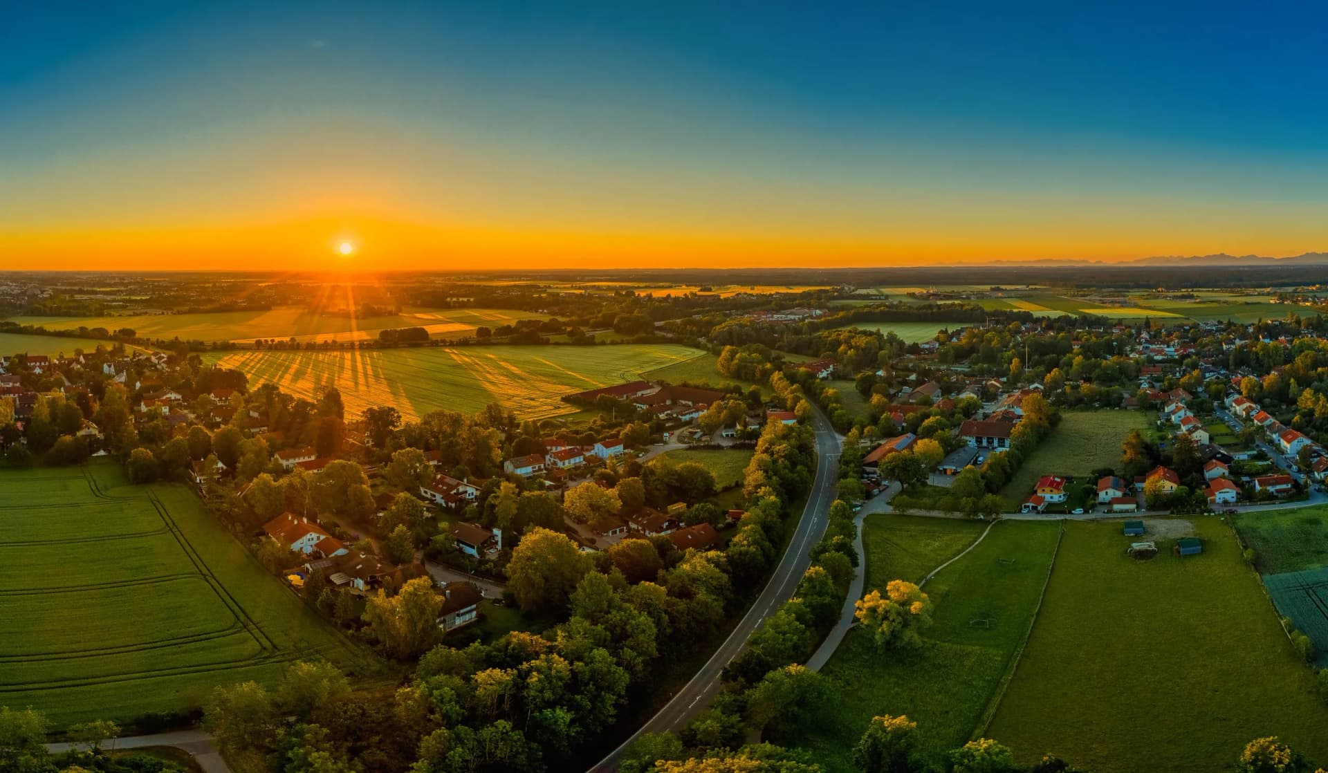 Aerial view of a village and green fields at sunset with distant mountains on the horizon.