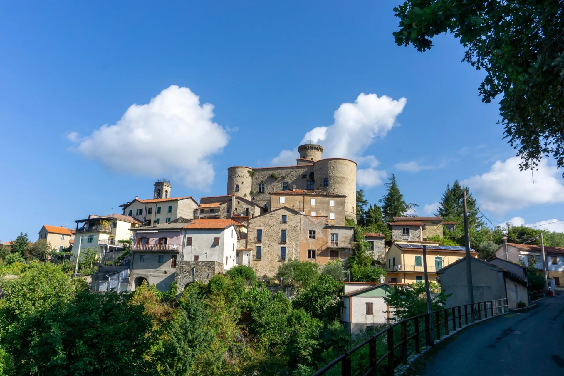 Hillside village with stone castle and terracotta roofs under a blue sky with clouds.