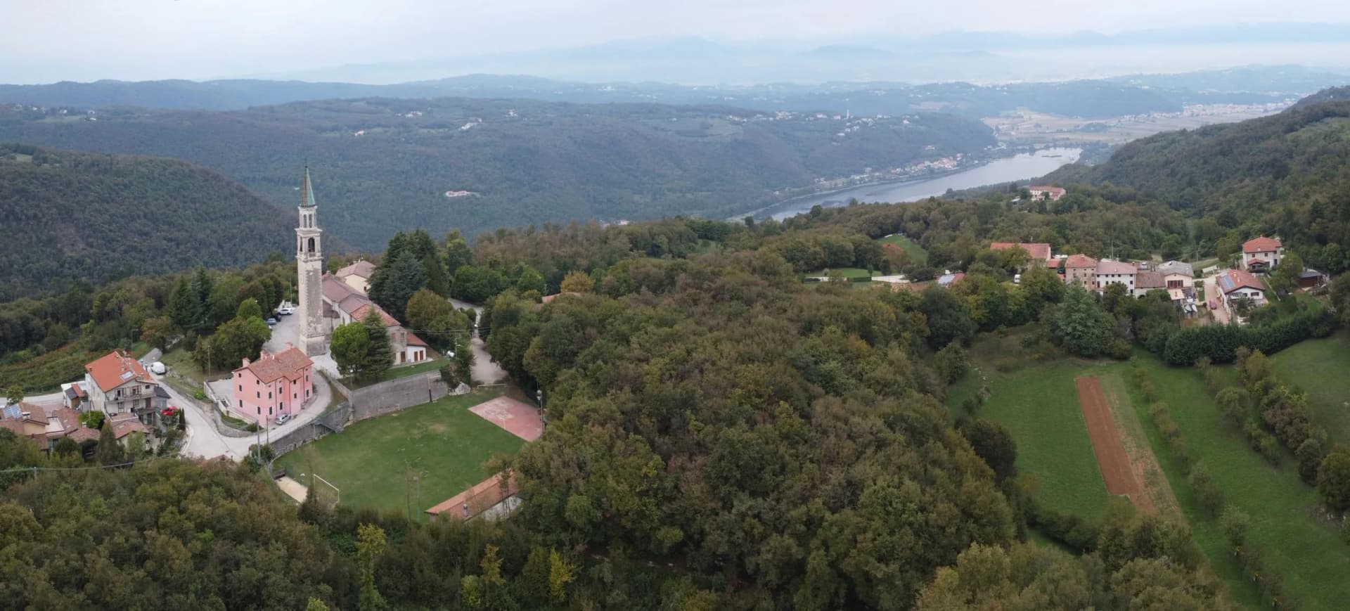 Village with church tower nestled in forested hills overlooking a distant river valley.