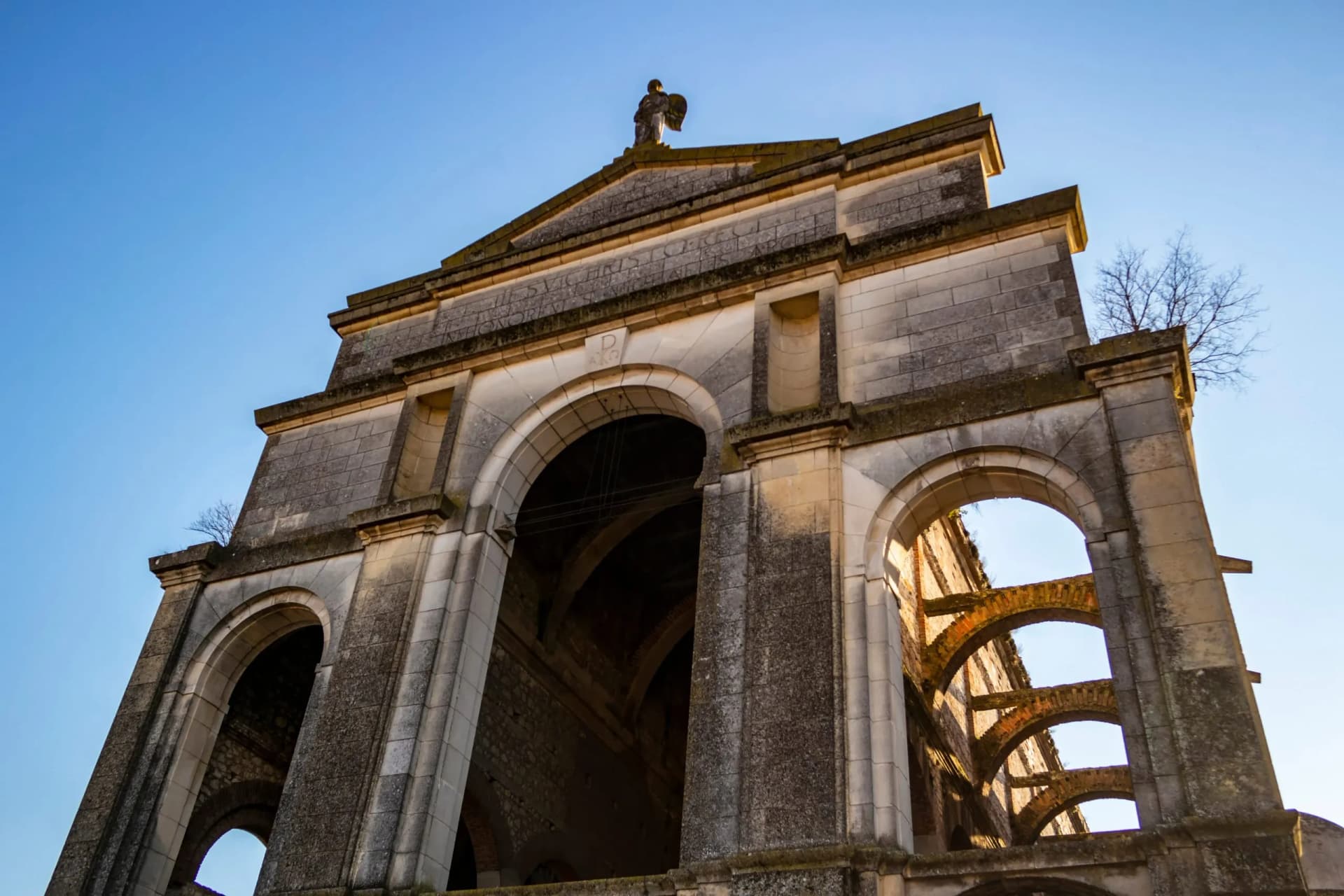 Ruined stone facade with large arches and a statue atop the pediment against a clear blue sky.