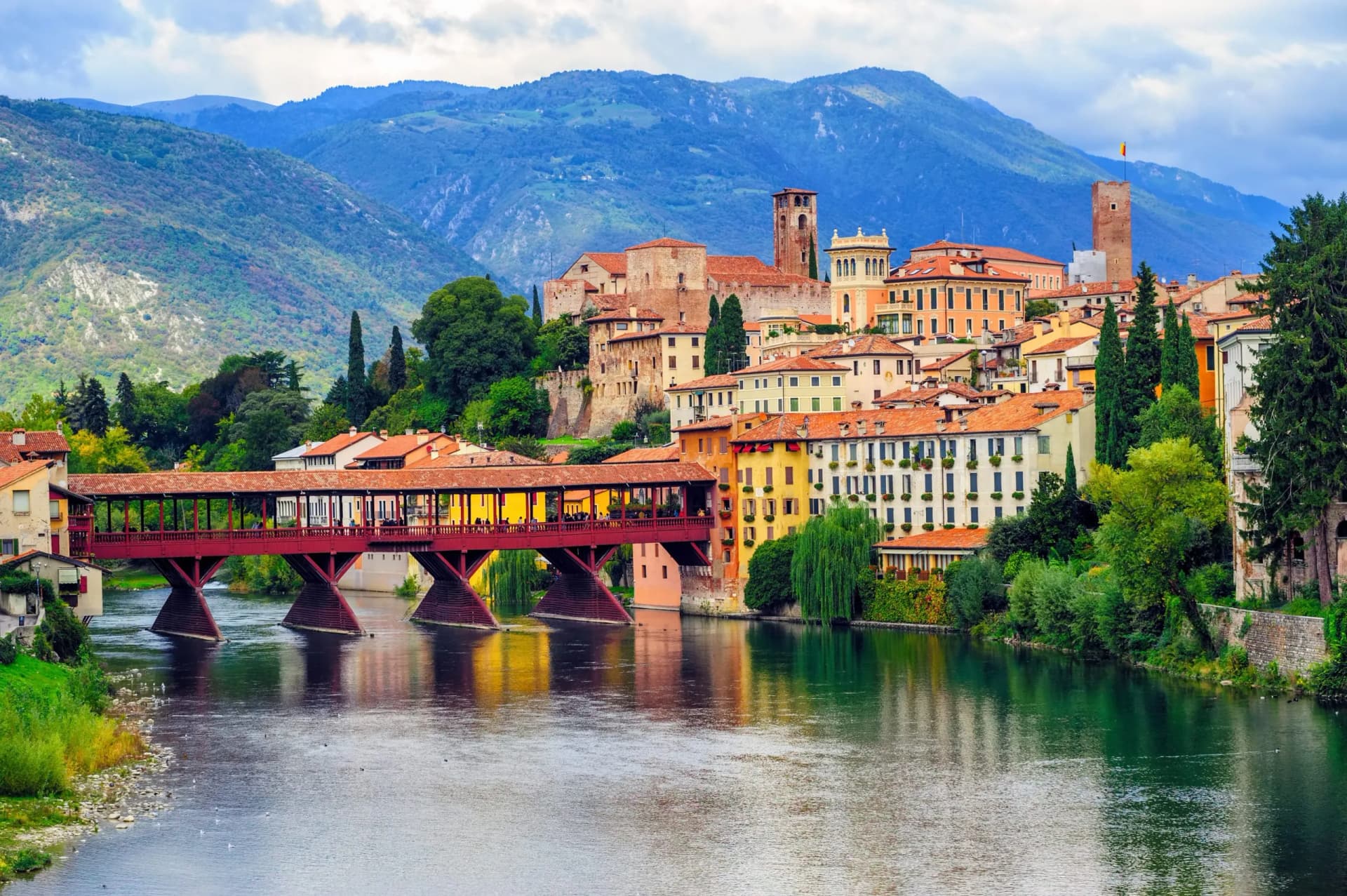 Red covered bridge over river in Vicenza with colorful buildings and blue mountains.