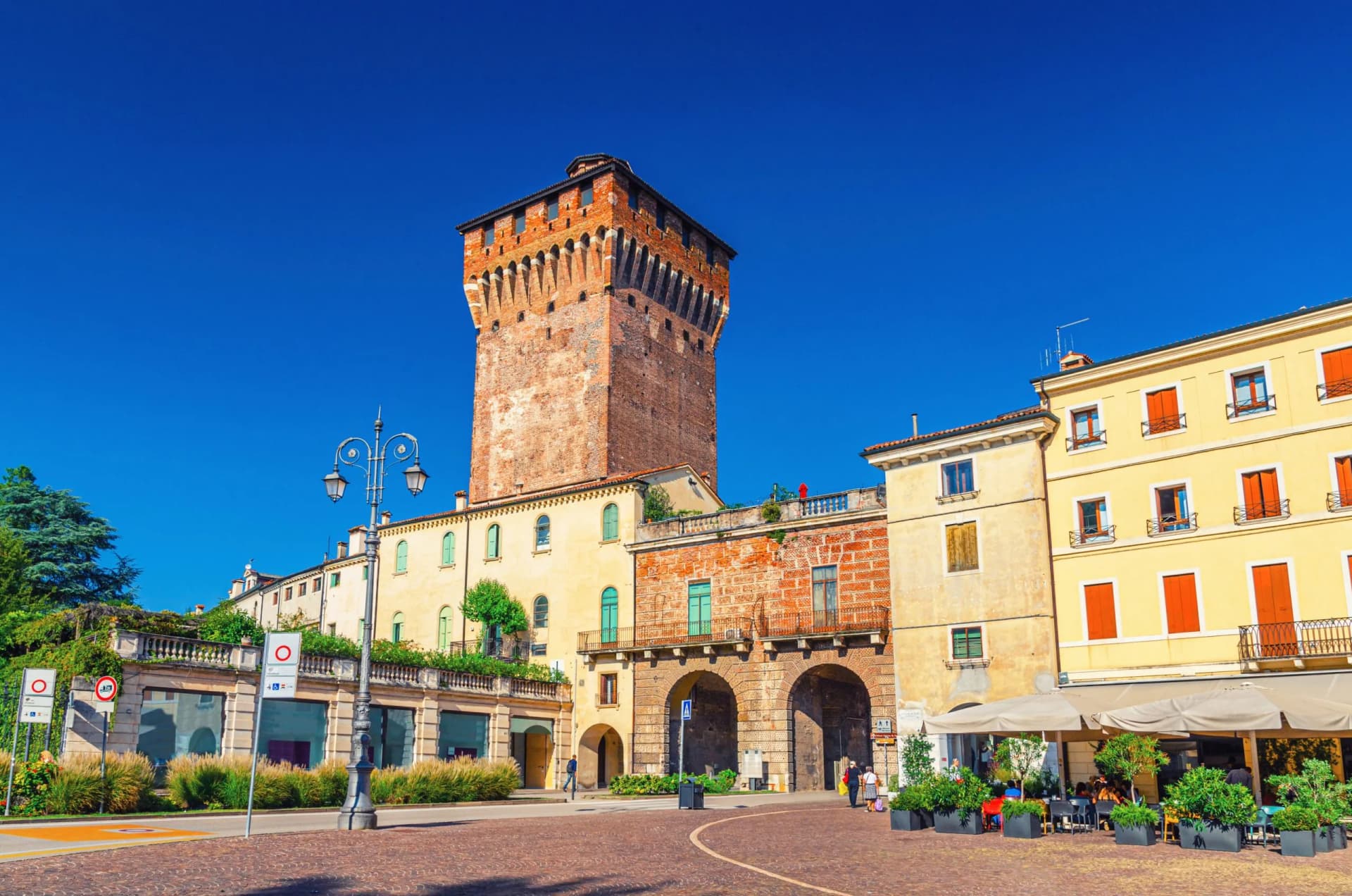 Historic brick tower next to yellow buildings with outdoor cafe seating in Vicenza square.