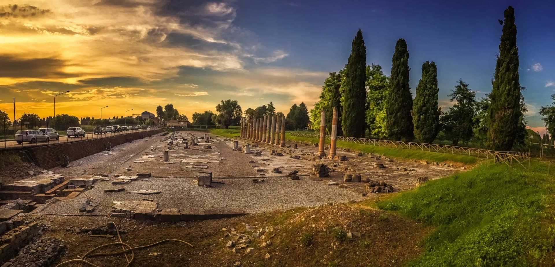 Roman ruins with standing columns, cypress trees, and traffic on a road at sunset in Aquileia.