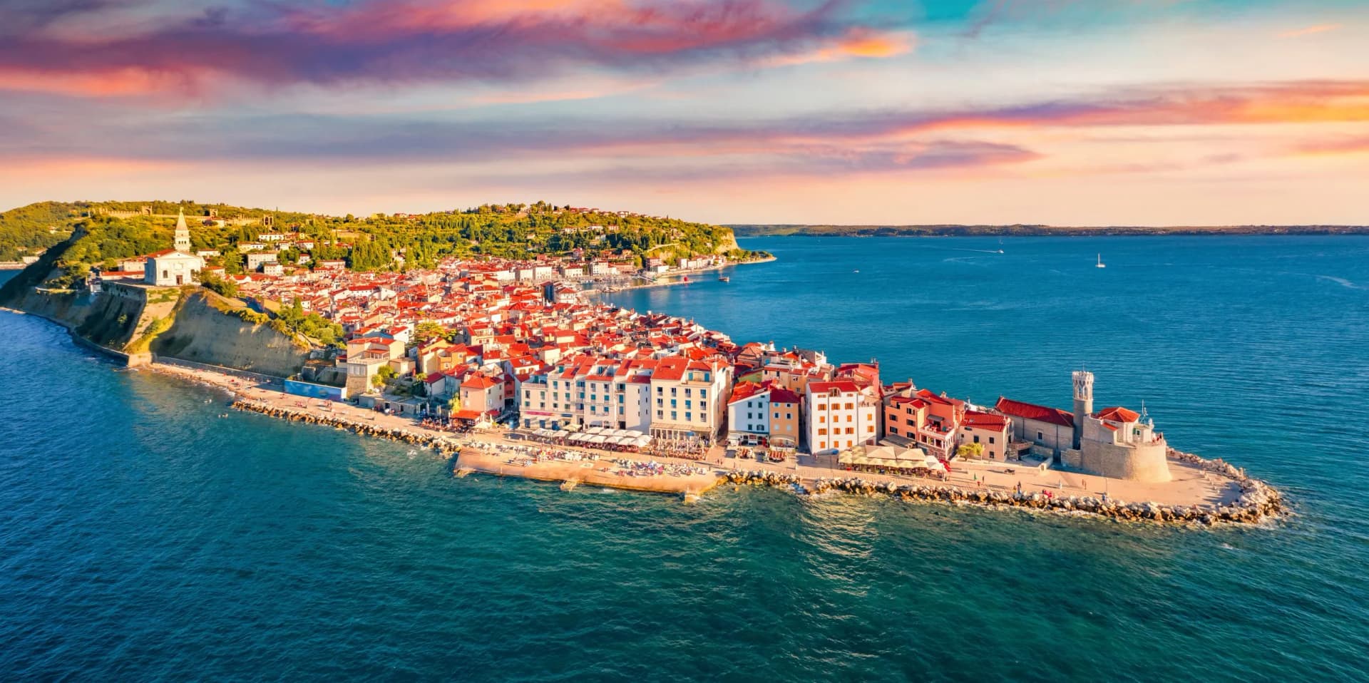 Coastal town with red-roofed buildings on a peninsula jutting into turquoise sea at sunset