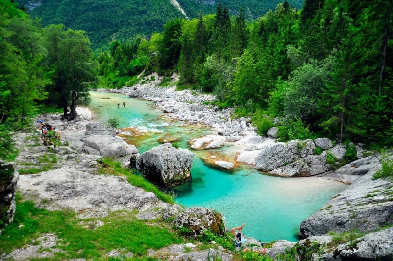 Hikers near turquoise river with clear water pools surrounded by lush green forest mountains.