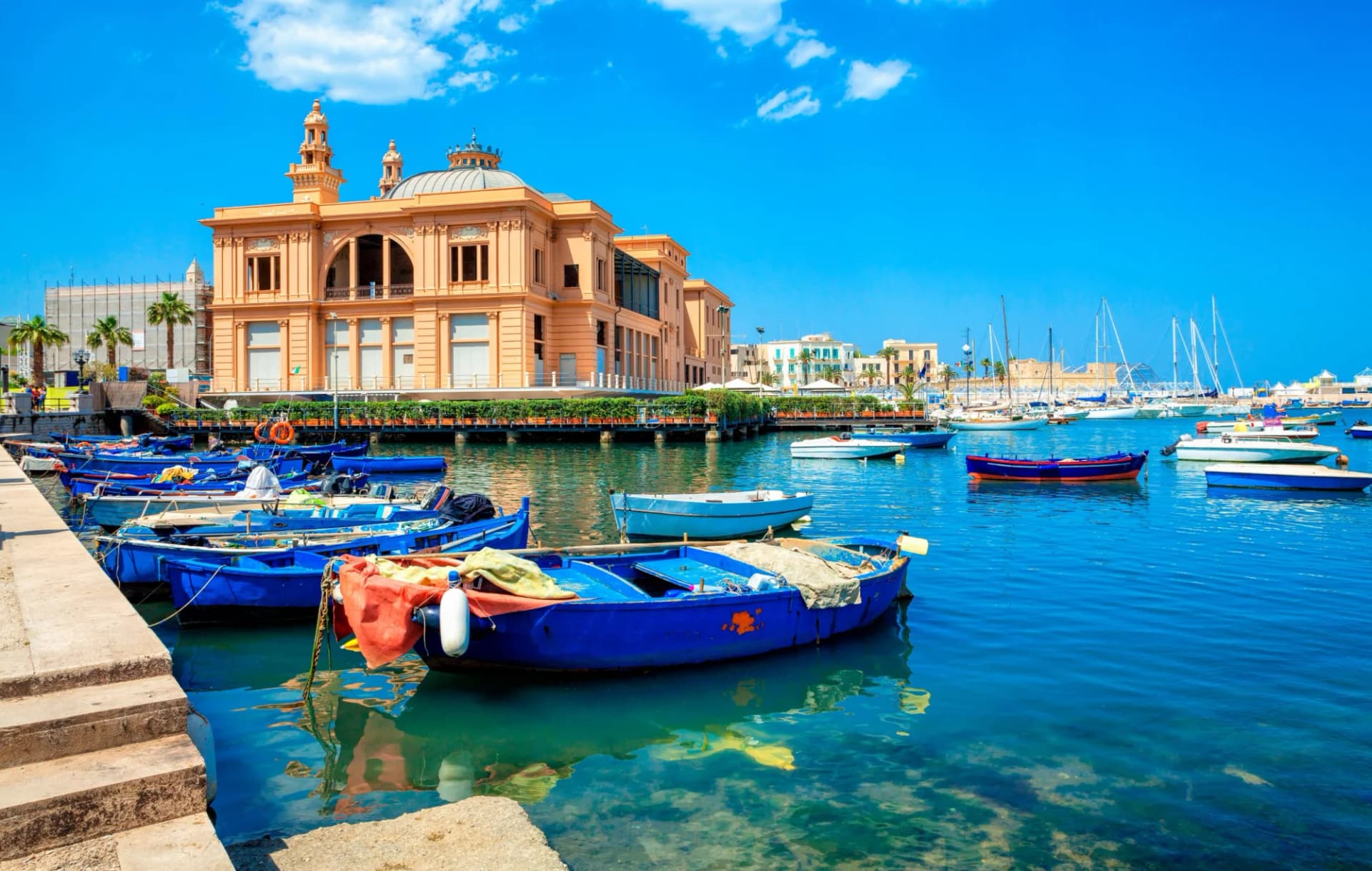 Blue fishing boats docked near a large ornate building in the Bari harbor under a bright blue sky.