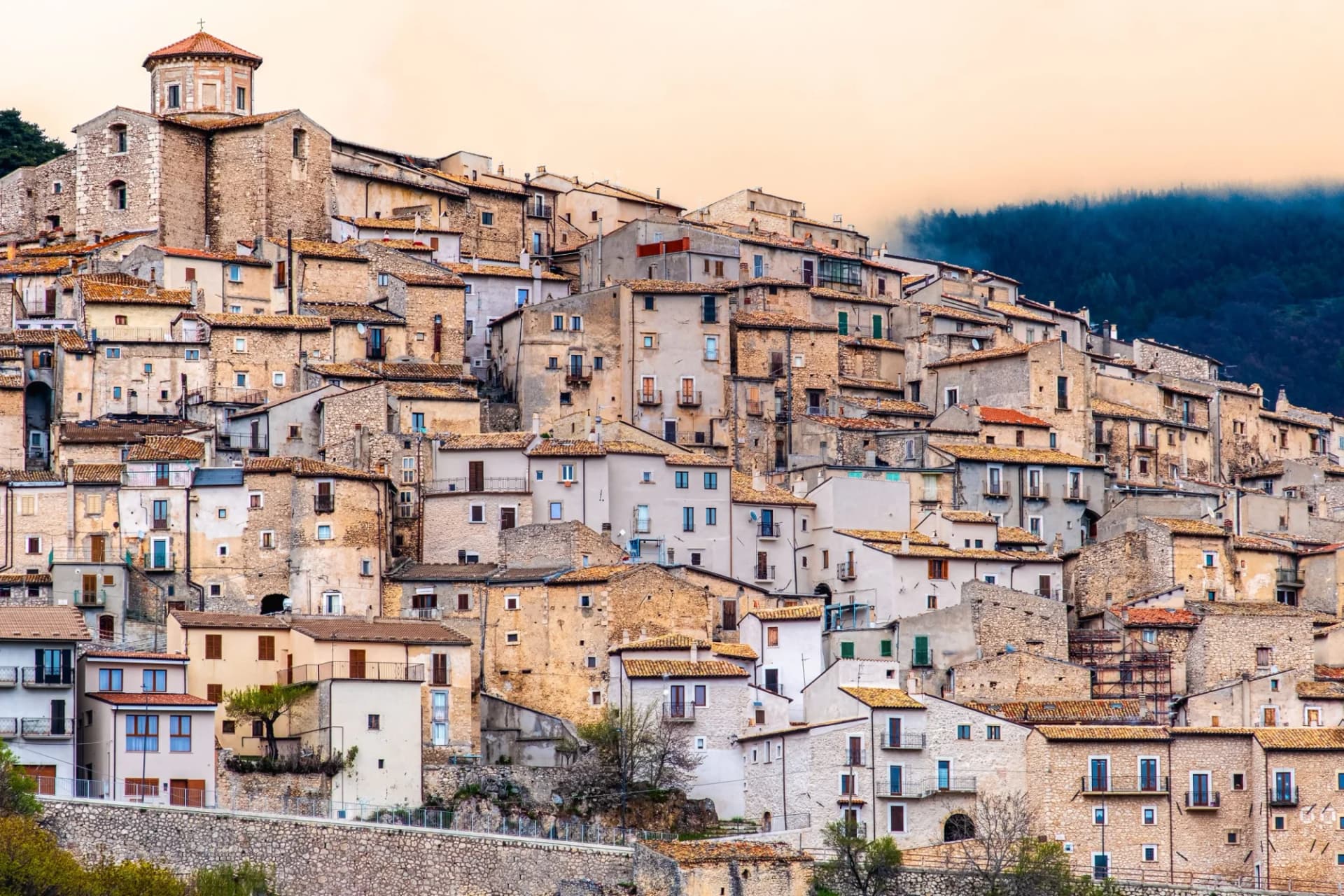 Hillside town with dense stone buildings and a church tower against a misty, forested mountain backdrop.