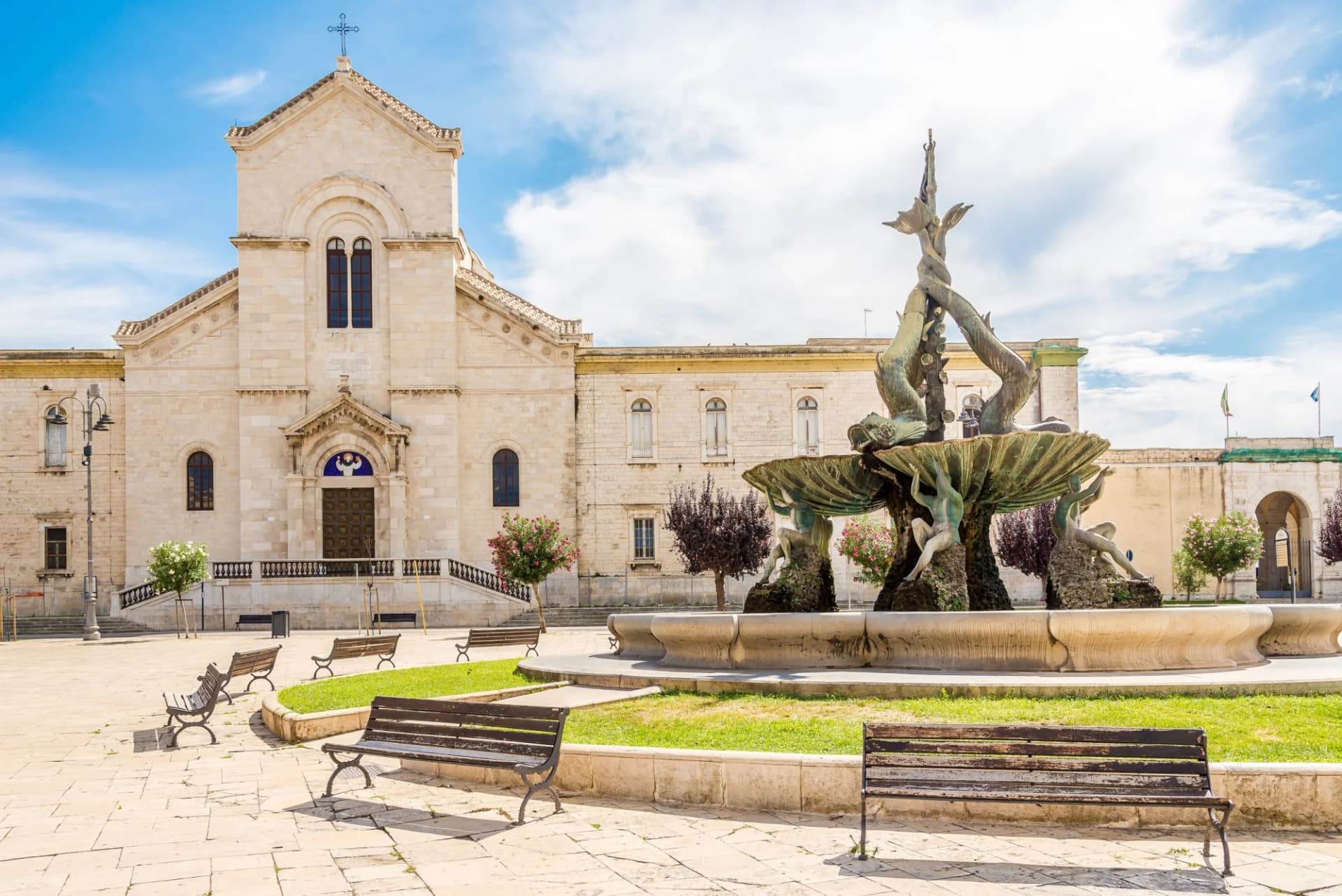 Stone church facade and bronze fountain in a sunny European town square with benches.