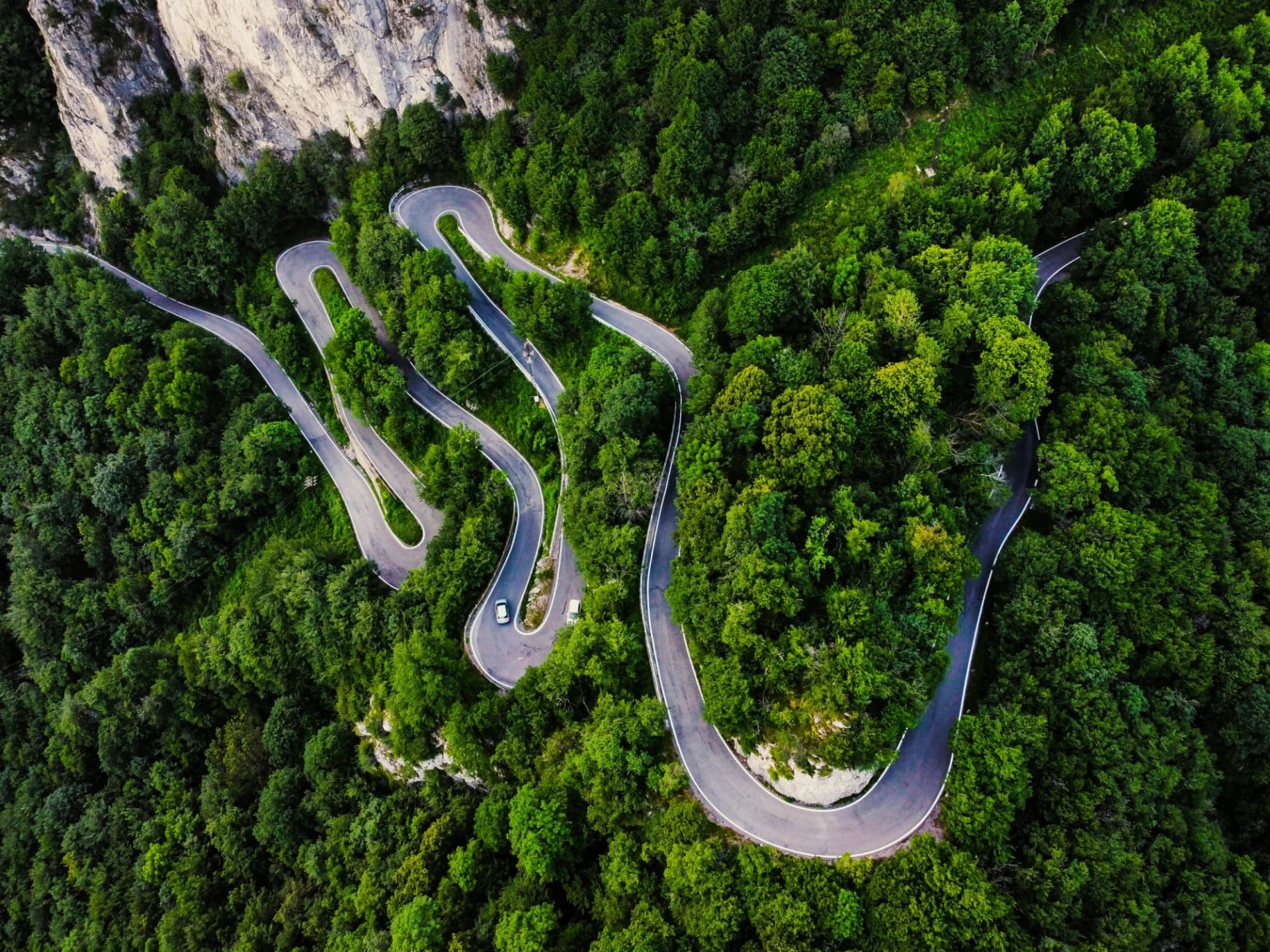 Winding mountain road with hairpin turns through dense green forest near a cliff face, Valsassina.