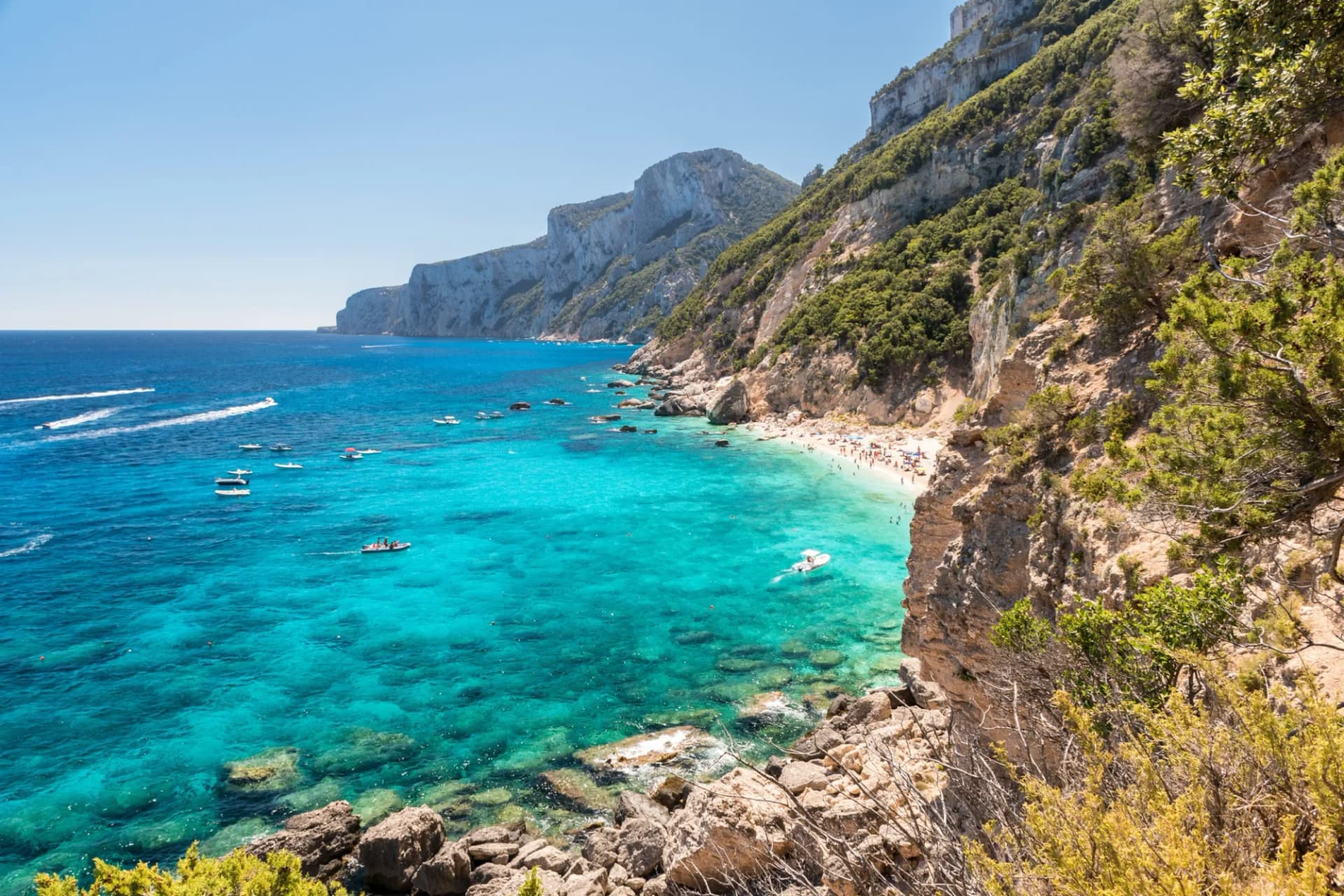 Boats on turquoise sea near beach cove with steep, vegetated cliffs, Gulf of Orosei.