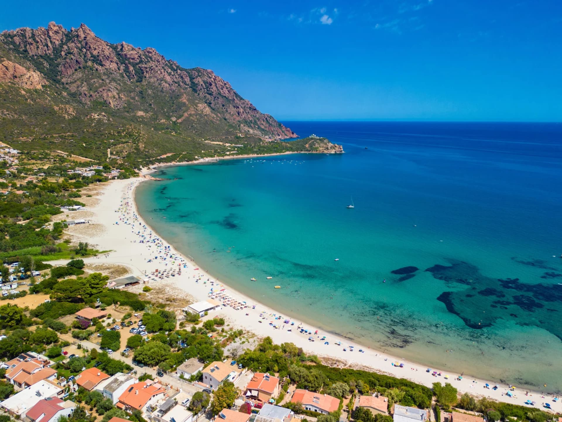 Aerial view of a crowded white sand beach, turquoise sea, and rocky mountains under a clear blue sky.