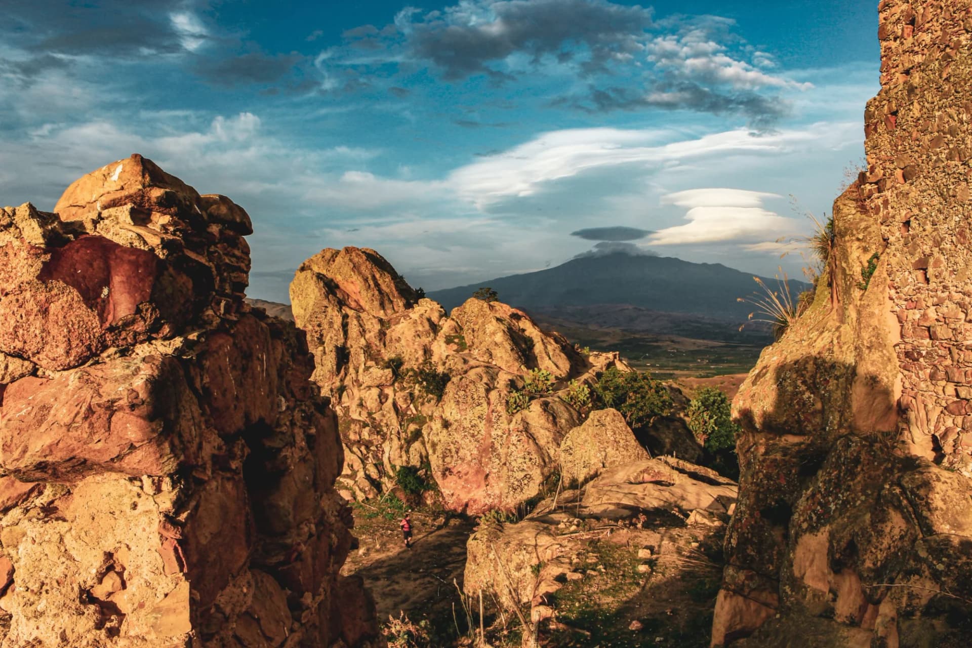 Rocky terrain with ancient stone walls overlooking a valley and distant mountain under a cloudy sky, Sicily Etna.
