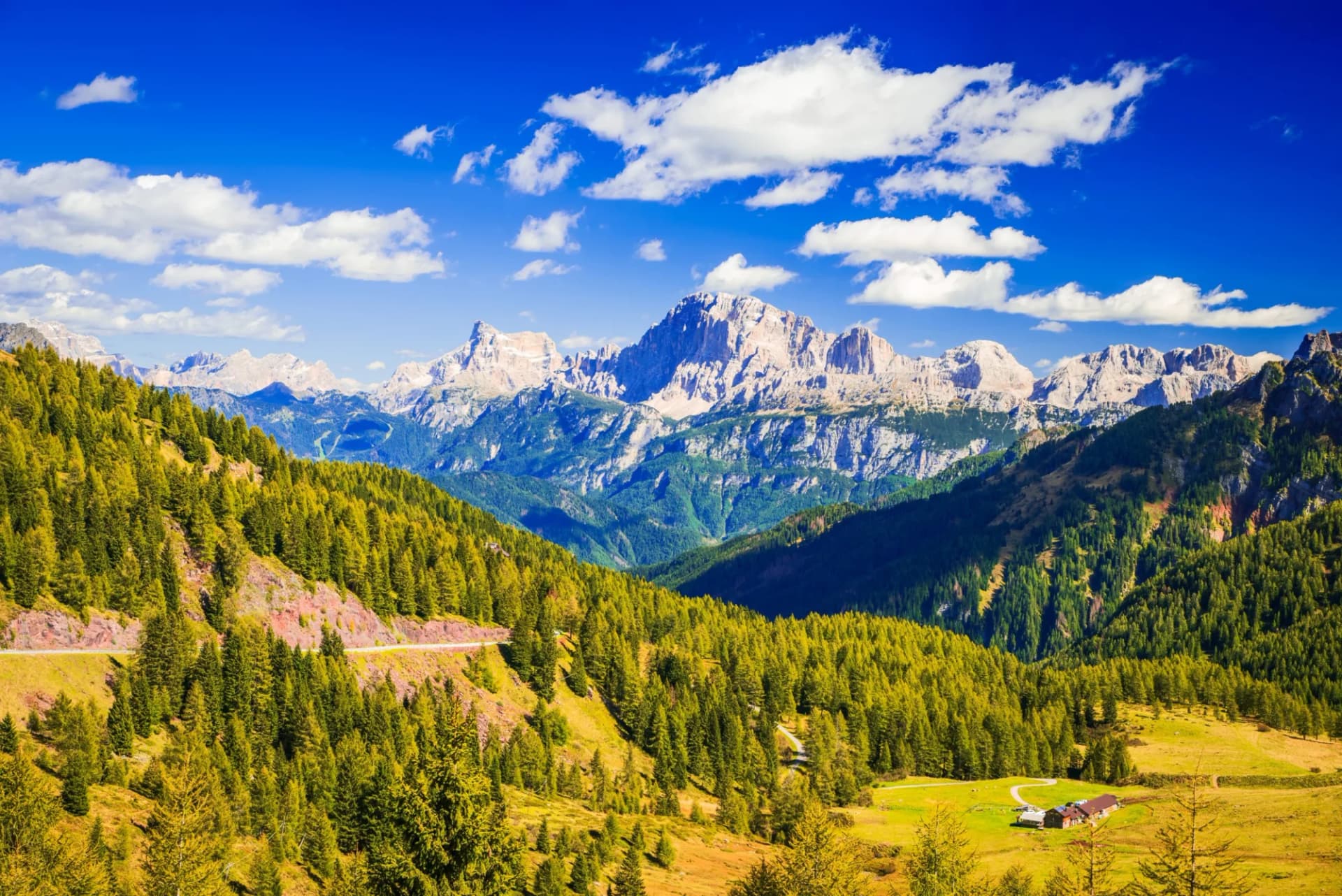 Alpine mountains with rocky peaks, green forests, and a bright blue sky in the Dolomites, Italy.