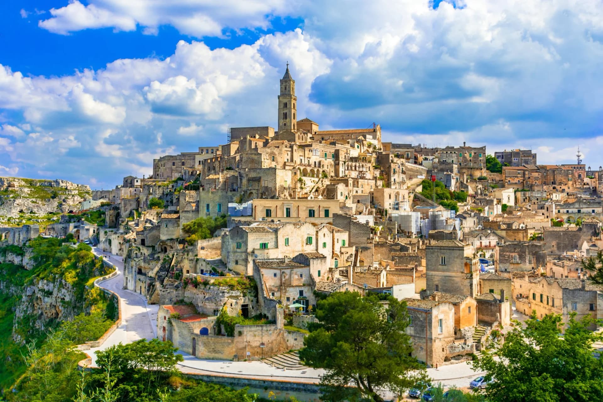 Historic stone city built into hillside with bell tower under bright blue, cloudy sky in Matera.