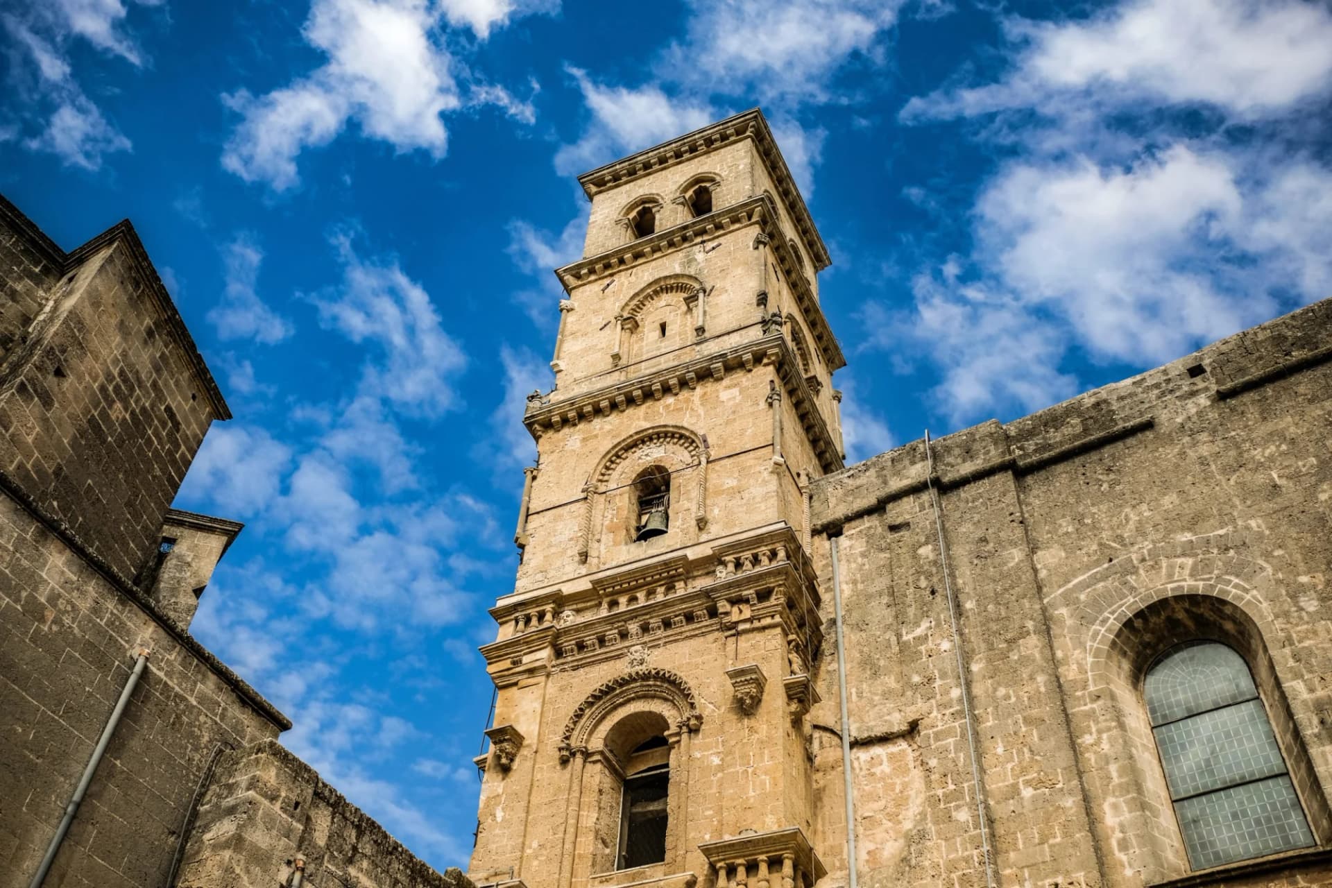 Tall stone bell tower of a historic church against a bright blue sky with white clouds.