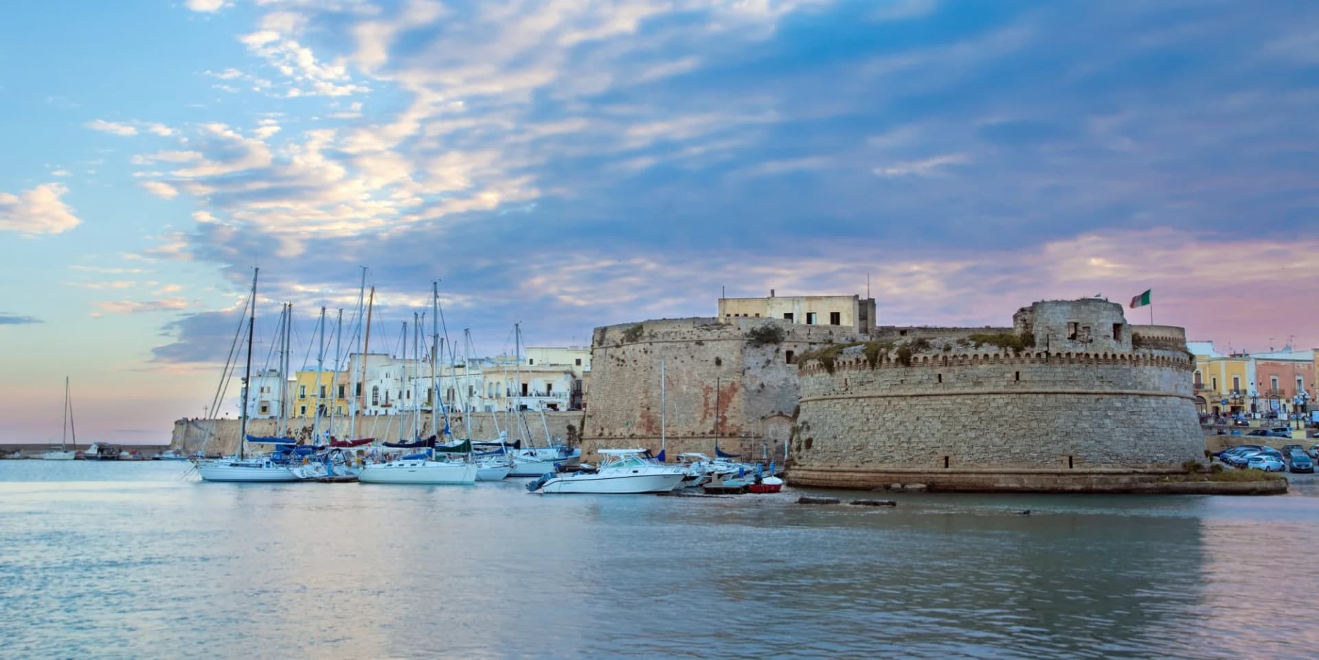 Sailboats docked near the historic stone castle of Gallipoli, Italy, under a dramatic sky.