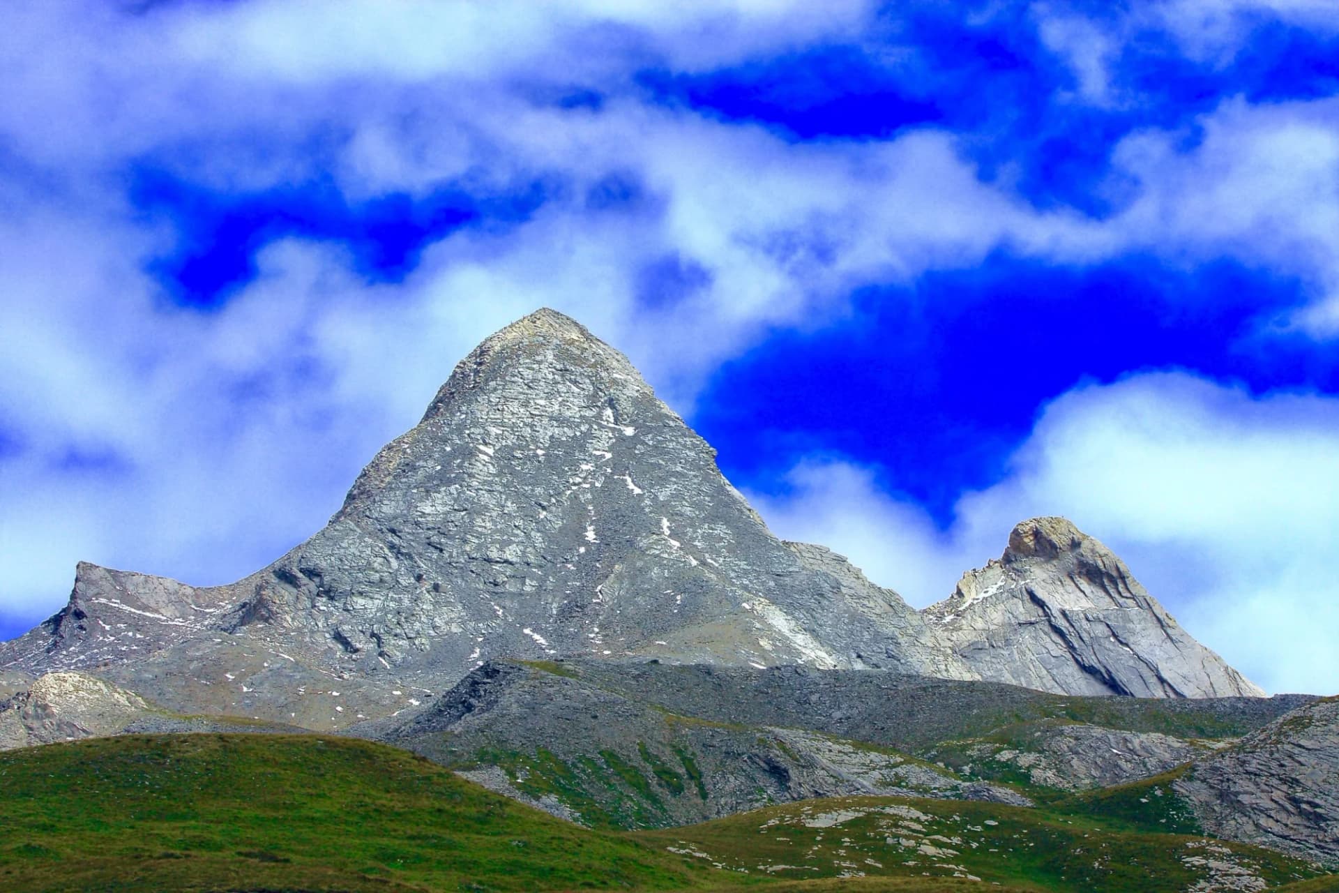 Rugged alpine mountain peaks rise above green foothills under a vibrant blue sky with white clouds.