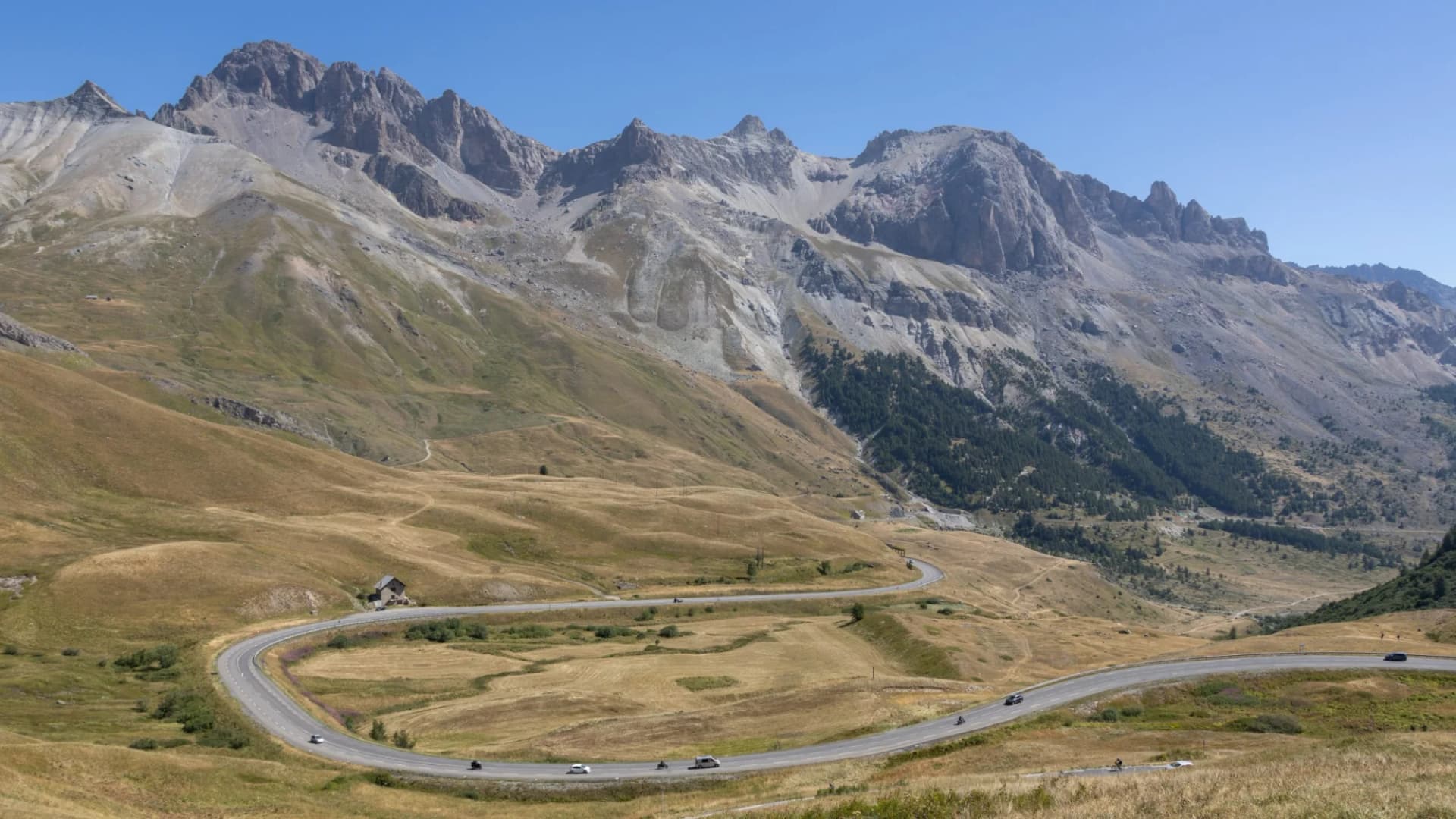 Winding mountain road with motorcyclists and cars ascending dry, grassy slopes near rocky peaks, Route du Col du Lautaret.