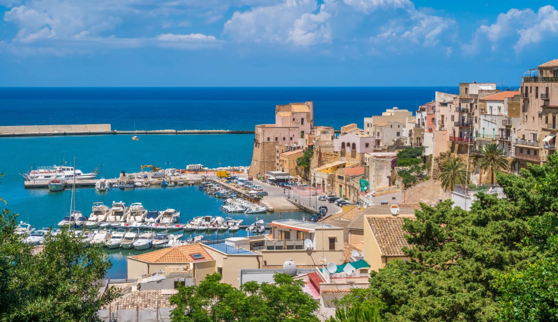 Boats docked in Castellammare del Golfo harbor with hillside town and blue Mediterranean sea.