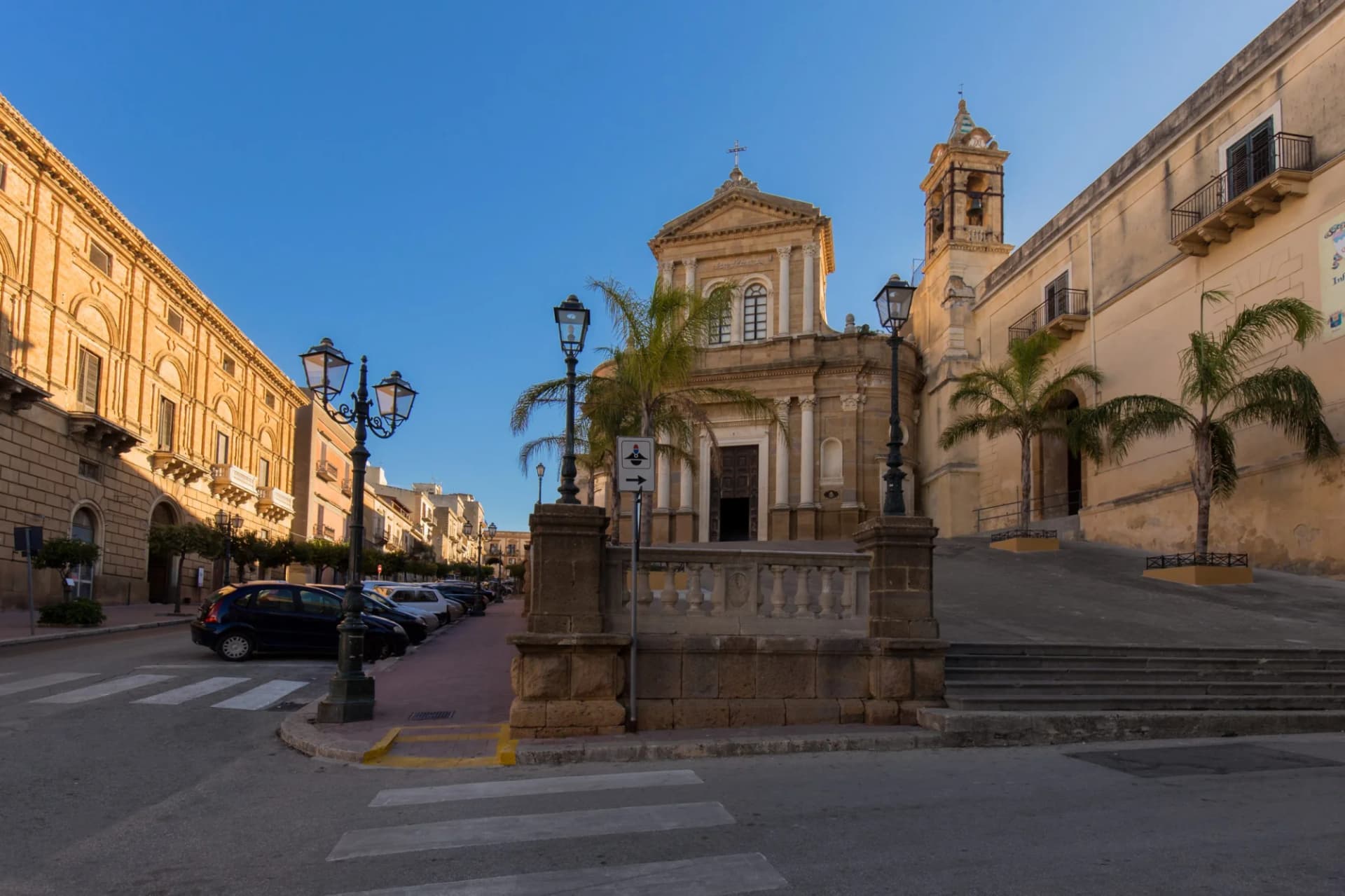 Baroque church facade with bell tower, palm trees, and street parking in a historic town square.