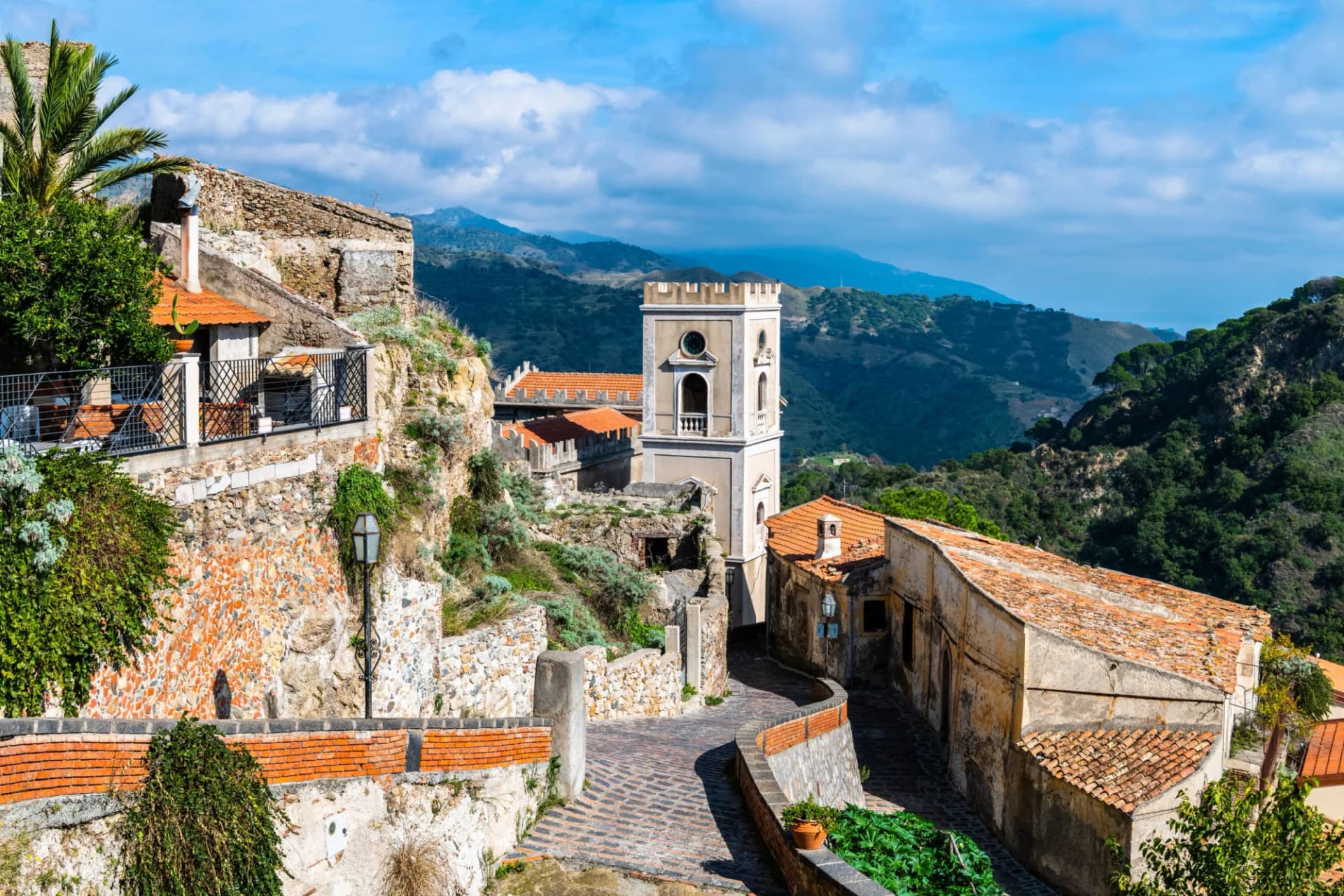 Winding cobblestone street in Savoca village, Sicily, with historic buildings and green mountains.