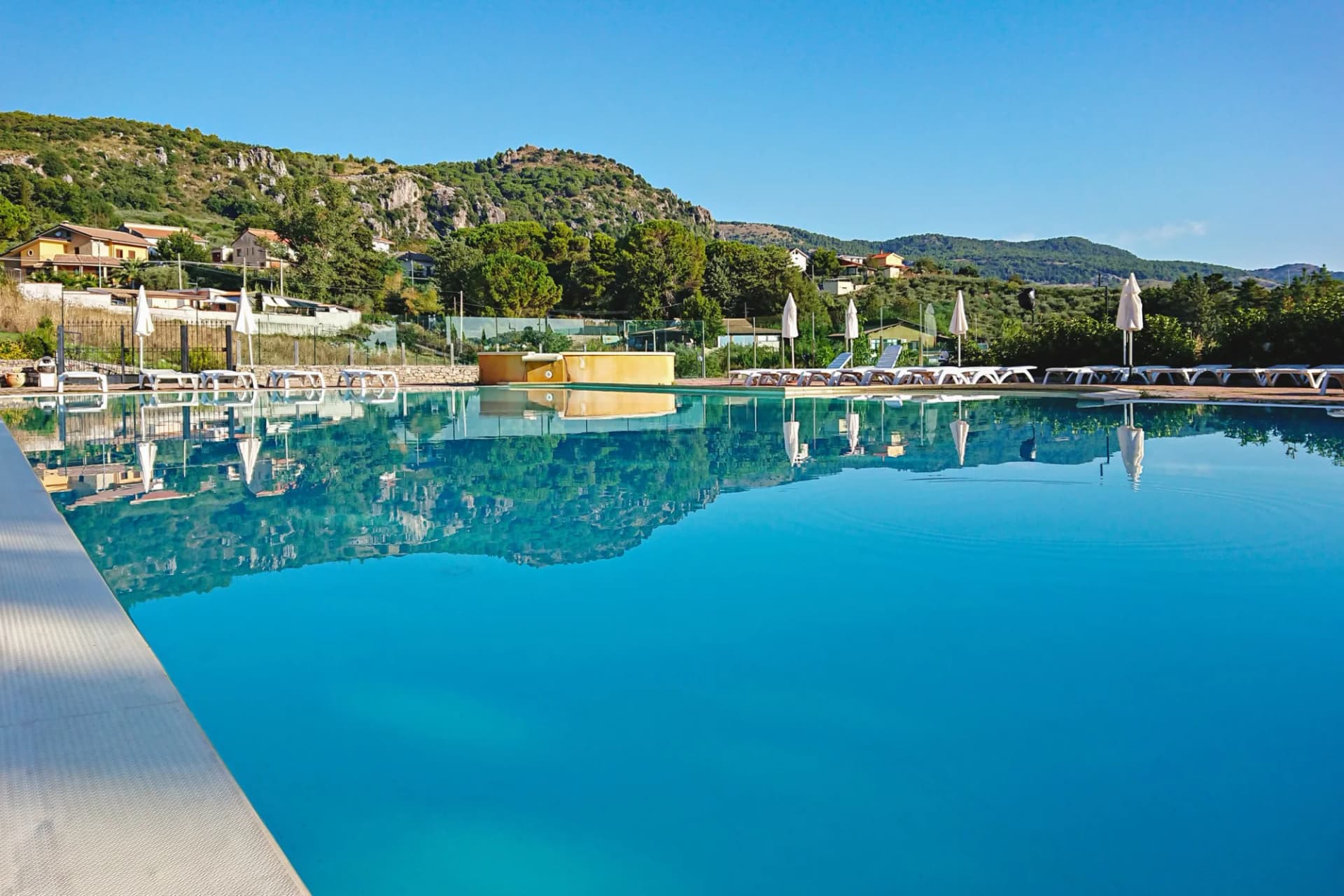 Swimming pool with lounge chairs reflecting on blue water, overlooking green hills in Piana degli Albanesi, Italy.