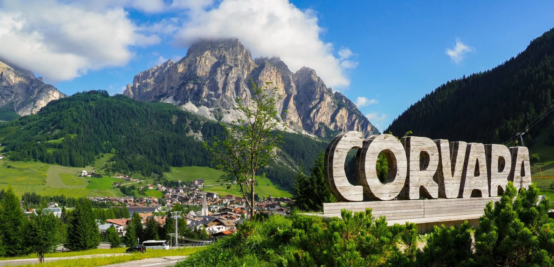 Large wooden "CORVARA" sign with alpine village and rocky mountains under blue sky.