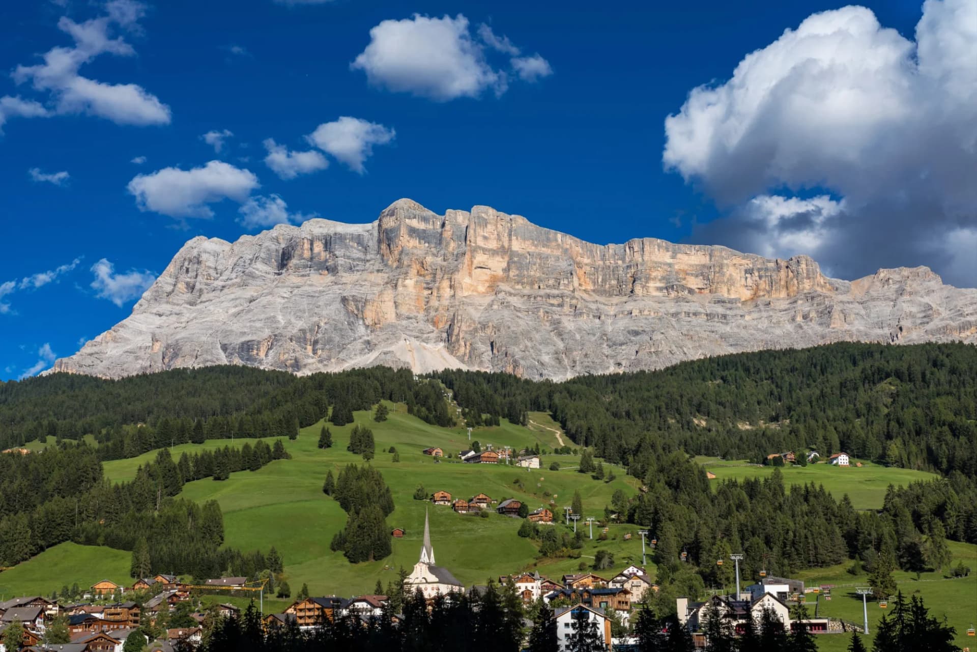 Alpine village nestled in green valley below massive gray mountain cliffs under blue sky.