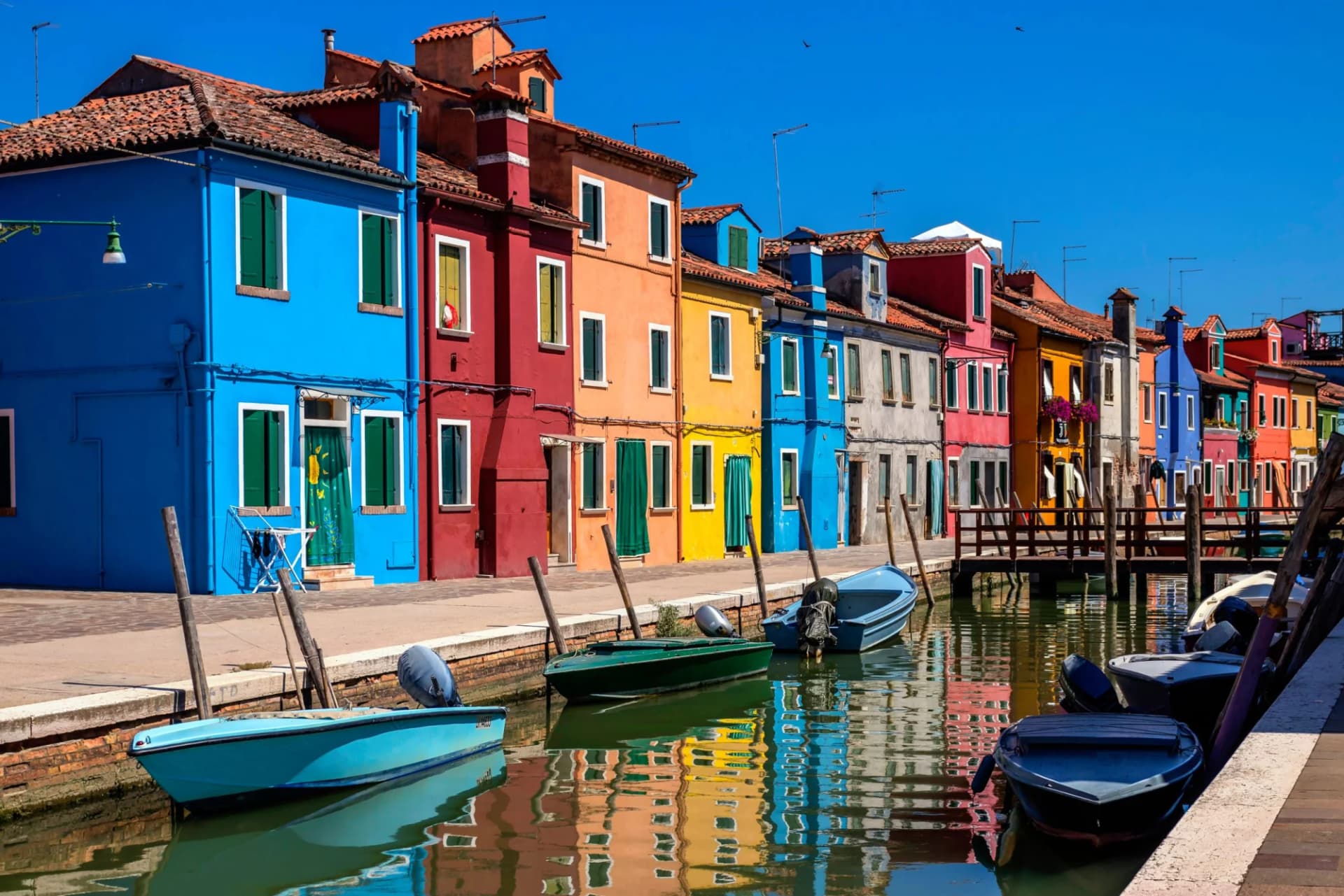 Colorful houses line canal with moored boats reflecting in water under clear blue sky.