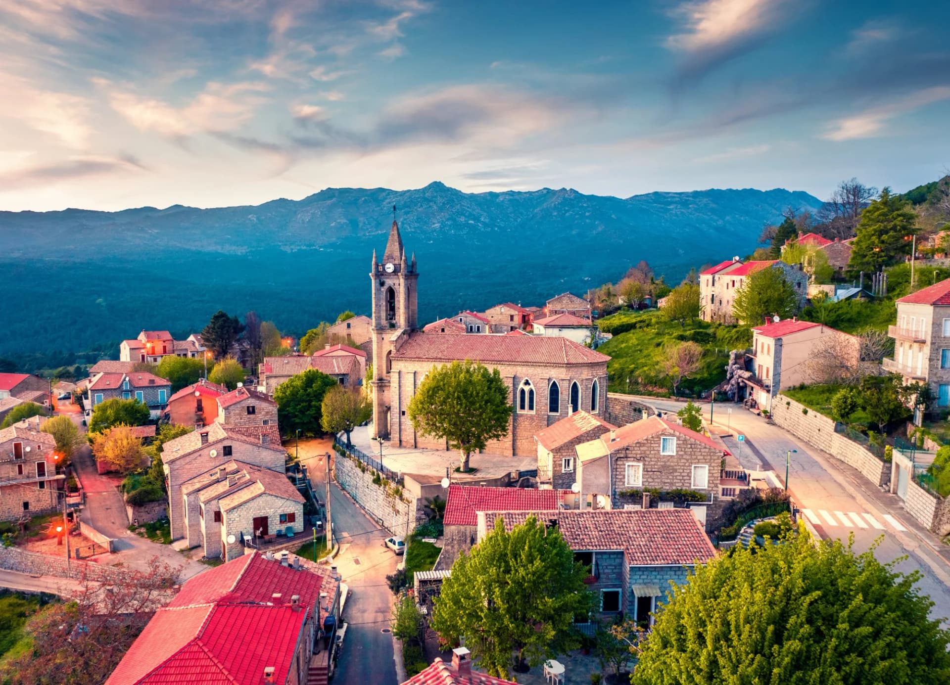 Village with stone houses and church tower nestled in green mountains under a dramatic sky.