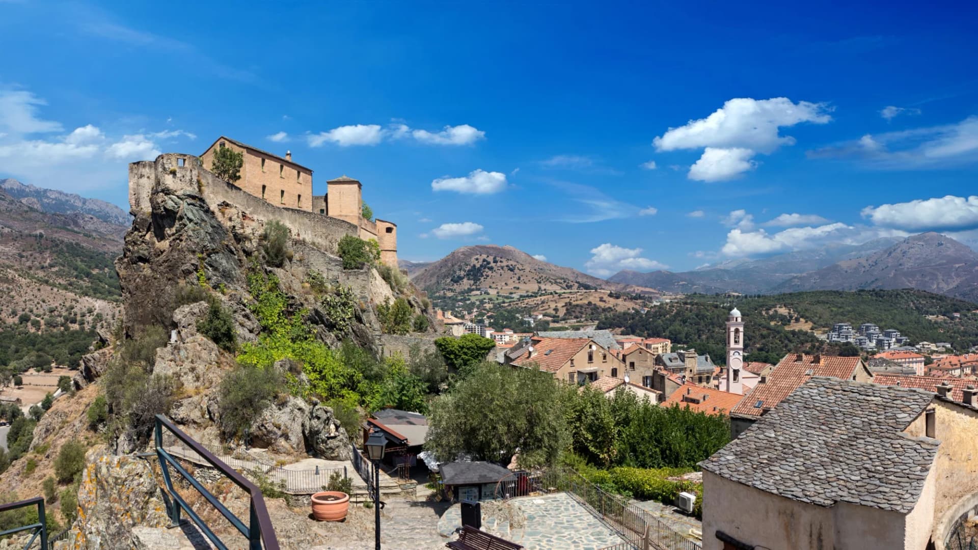 Fortress on rocky outcrop overlooking town with terracotta roofs and mountains under blue sky.