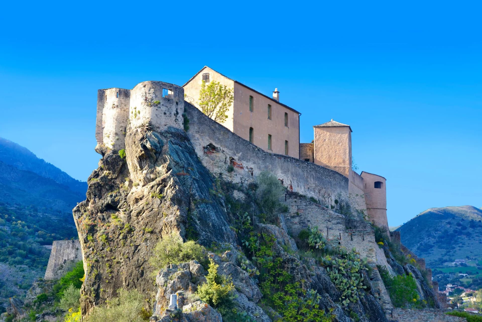 Citadel and buildings perched atop a steep, rocky outcrop under a clear blue sky in Corte, Corsica, France.